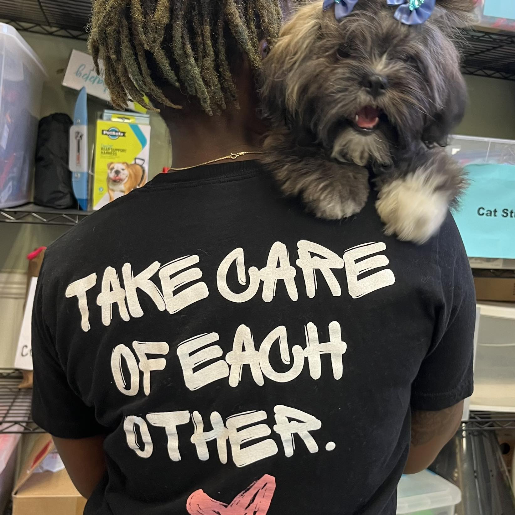 Person with dreadlocks wearing a black T-shirt with white text, holding a small dog with bows on its head.