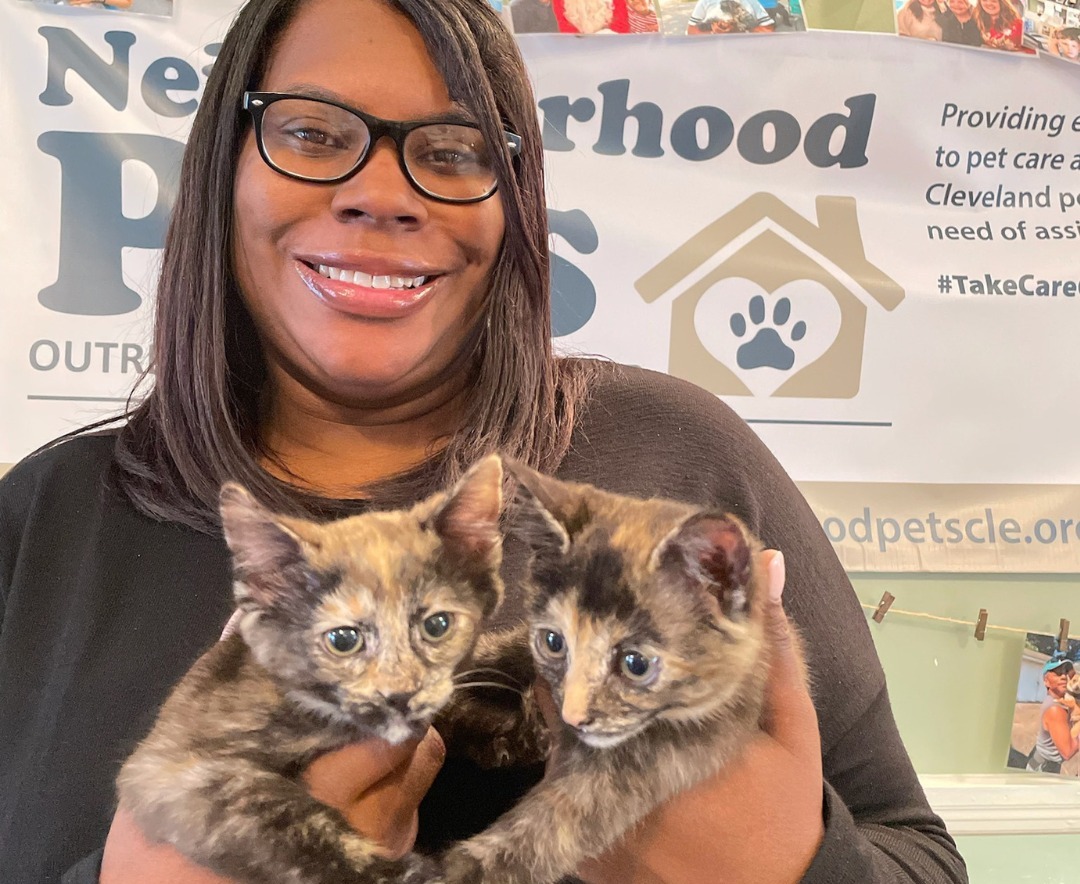 Woman holding two kittens, smiling, in front of a banner with a house and paw print logo.