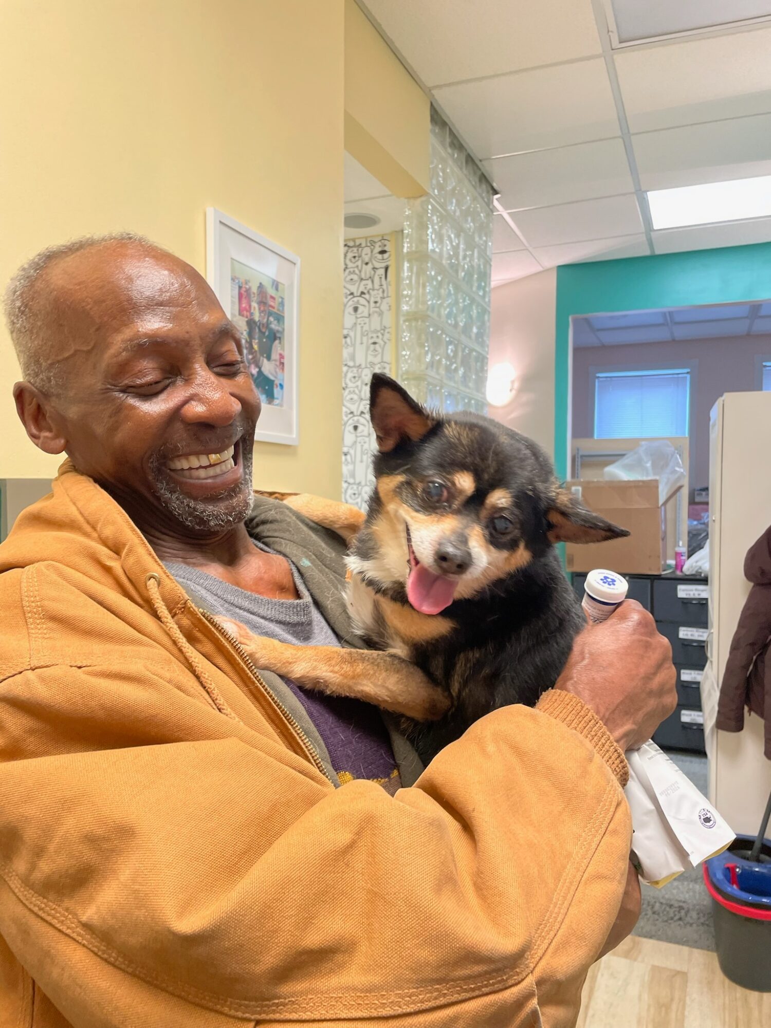 Elderly person smiling and holding a small dog indoors, with a colorful background and framed picture on the wall.