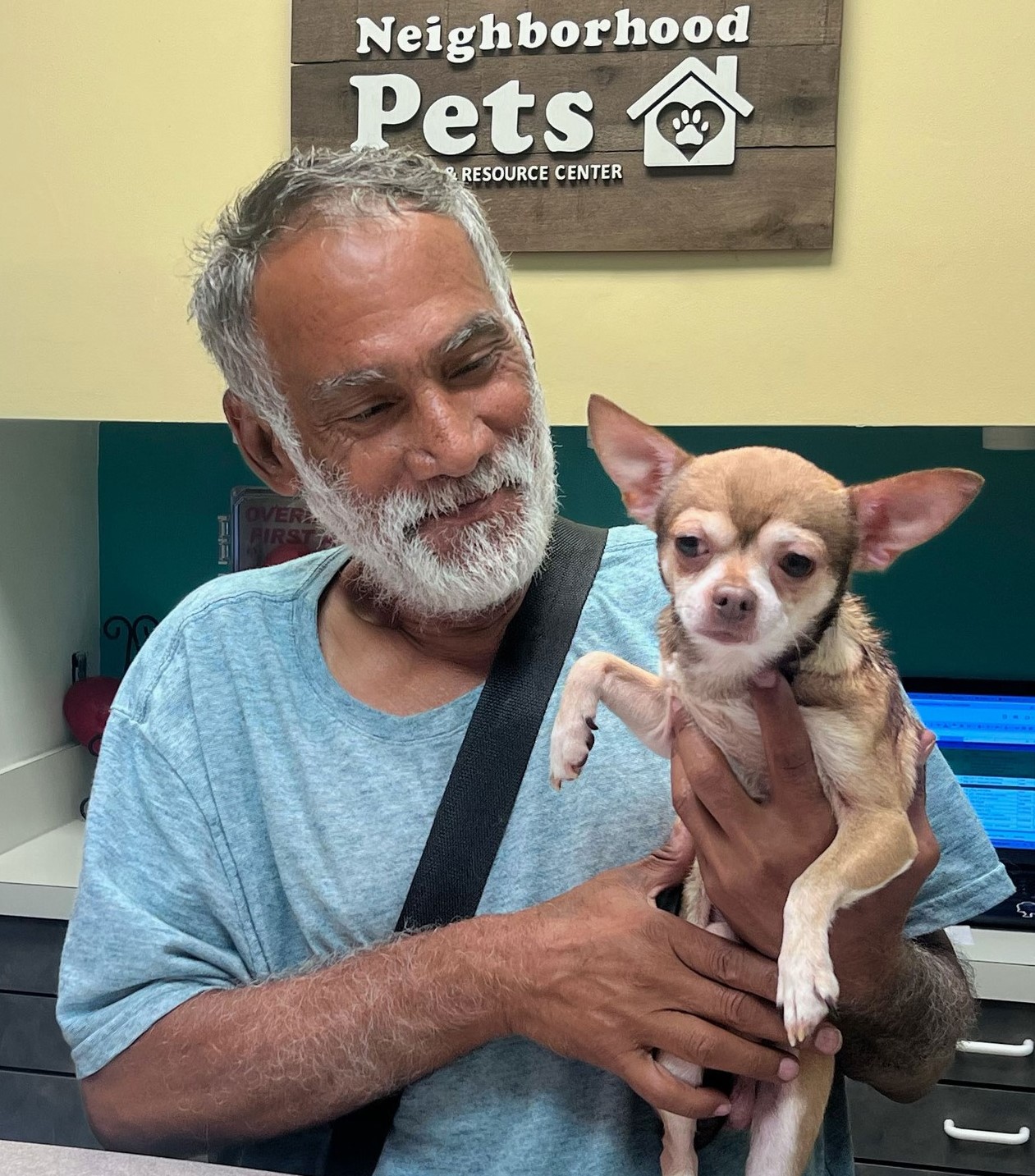 Older man with gray hair and beard holding a small dog with large ears, standing in front of a sign that reads 'Neighborhood Pets'.