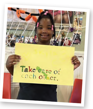 Young girl holding a yellow sign with colorful text, standing in front of a sports net, smiling. Word on sign: 'Take care of each other.'