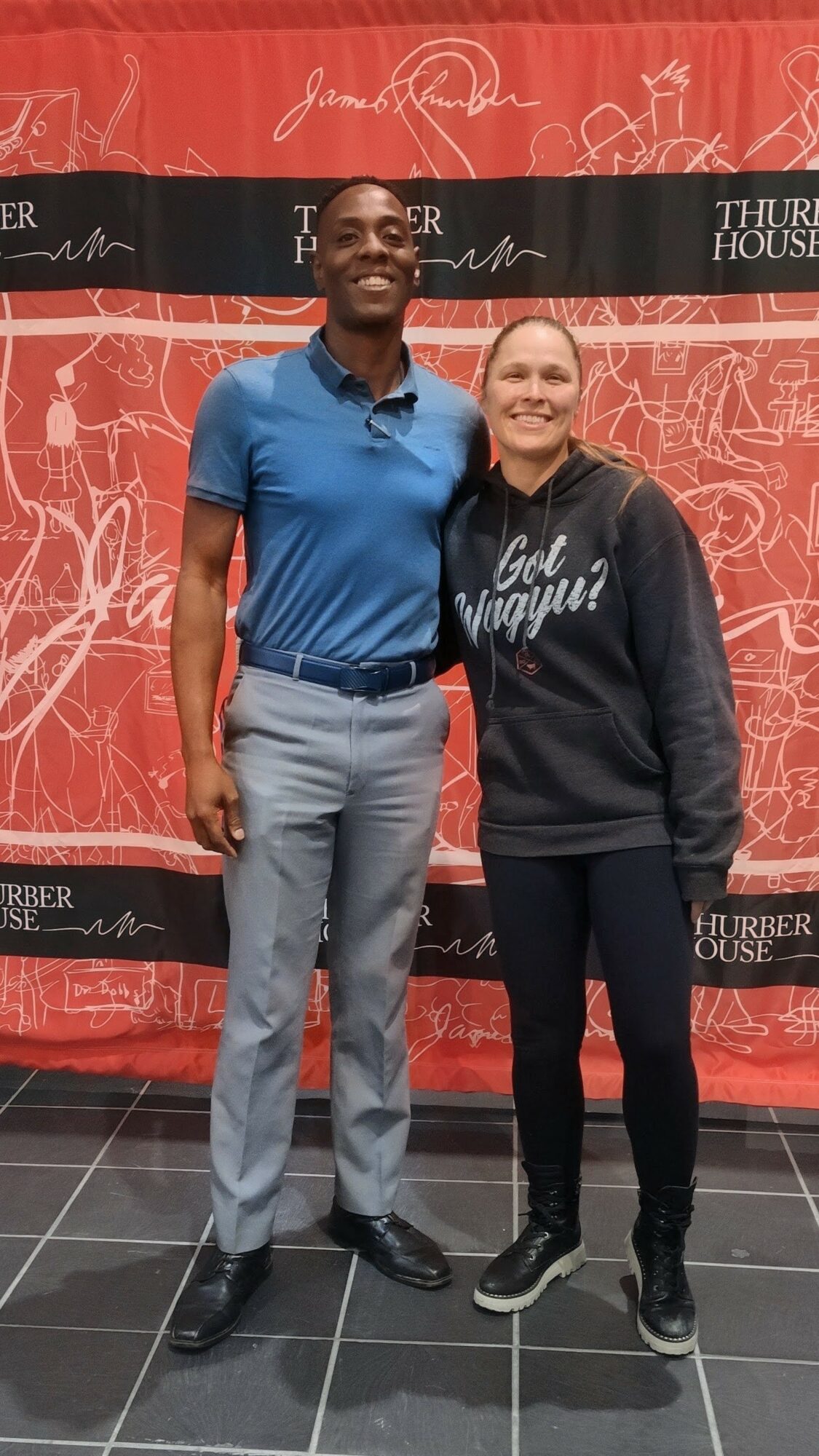 Two people standing together, smiling, in front of a red backdrop with signatures and text, one wearing a blue shirt and the other a dark hoodie.