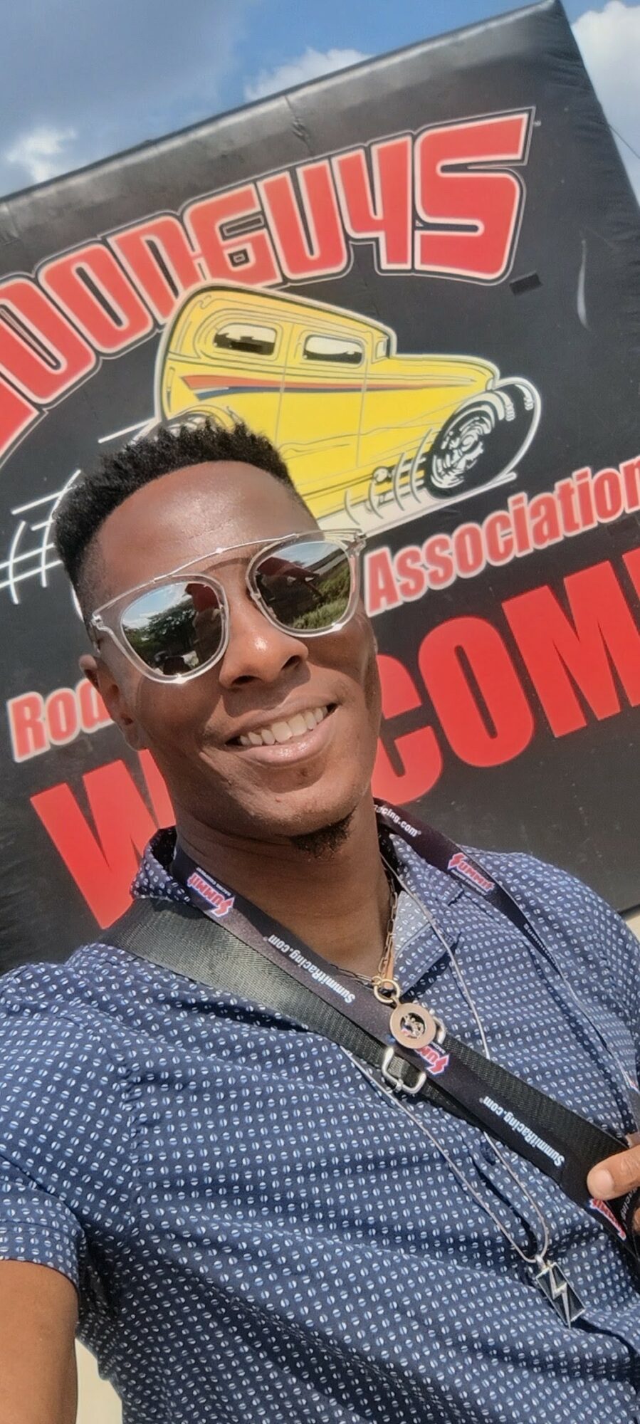 Person wearing sunglasses smiling in front of a sign with a yellow car illustration and red text.