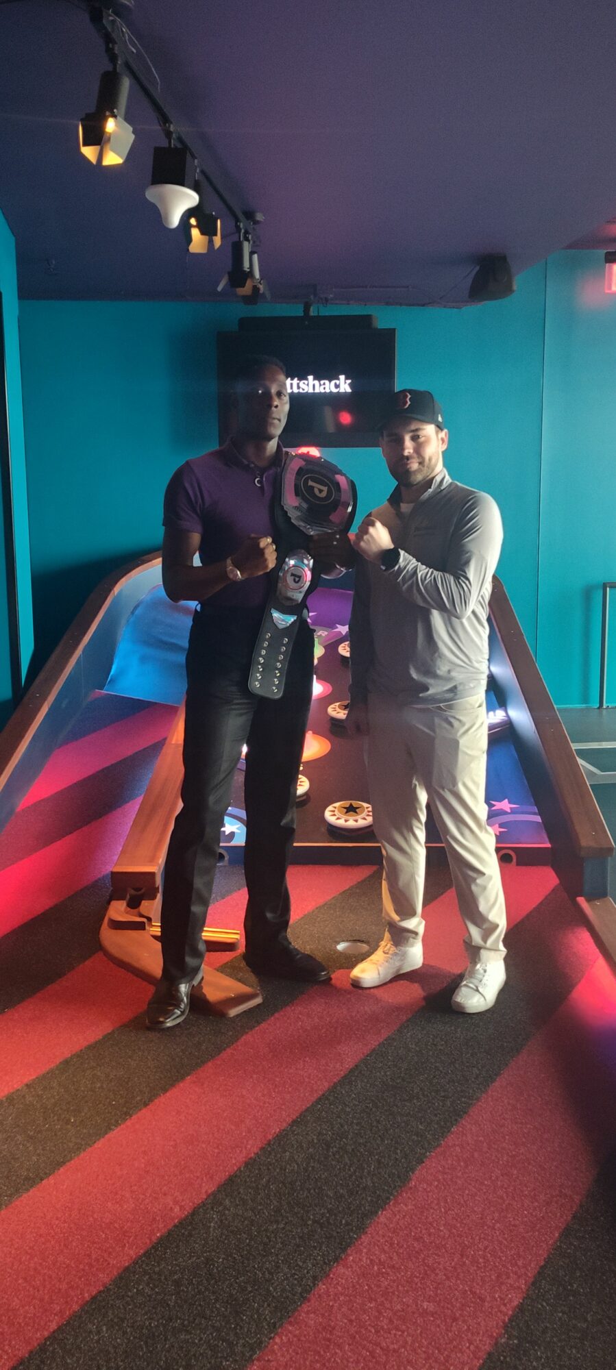 Two people stand in a room with colorful lighting, one in a purple jacket and the other in a gray jacket, near a pool table.