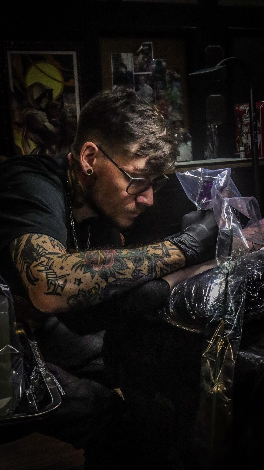 Person with glasses and tattoos working on a tattoo in a dimly lit studio, surrounded by artwork and equipment.