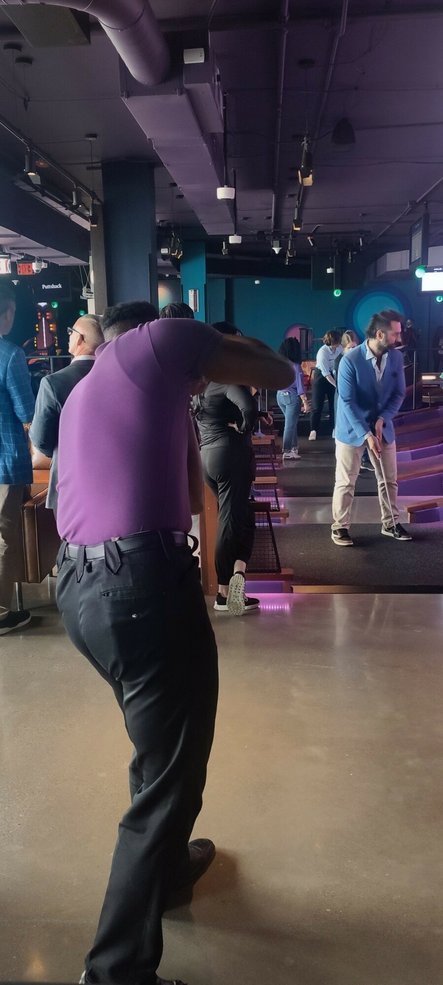 Person in purple shirt standing in a crowded indoor space with people and screens, facing away from the camera.