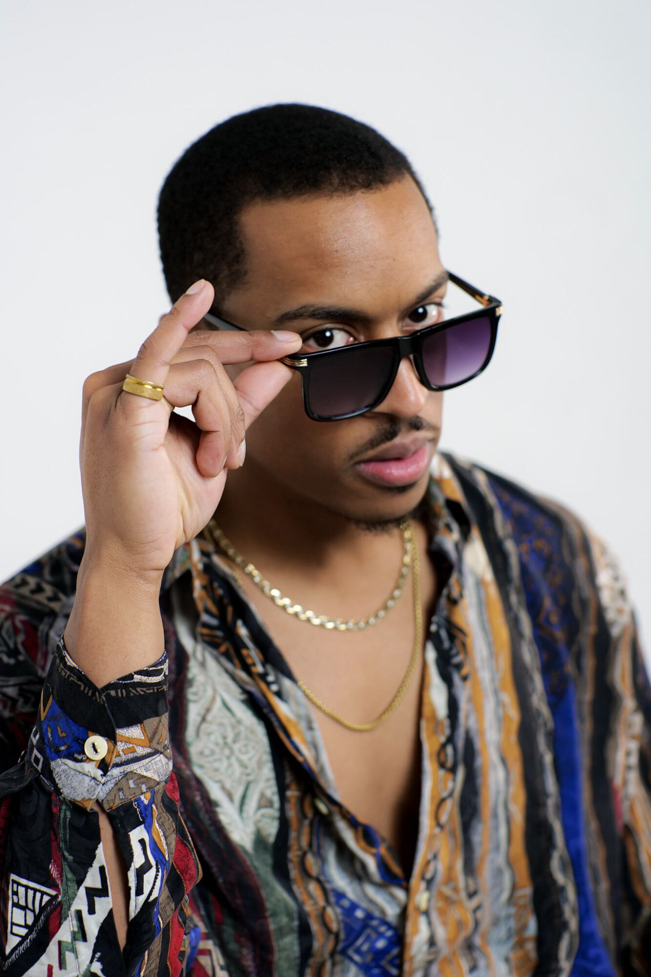 Person with short hair, wearing sunglasses, a patterned shirt, and a pearl necklace, looking at the camera.