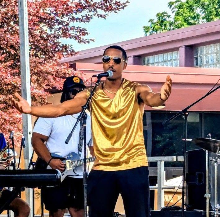 Man wearing sunglasses and a gold sleeveless shirt singing into a microphone outdoors, with a person playing guitar behind him.