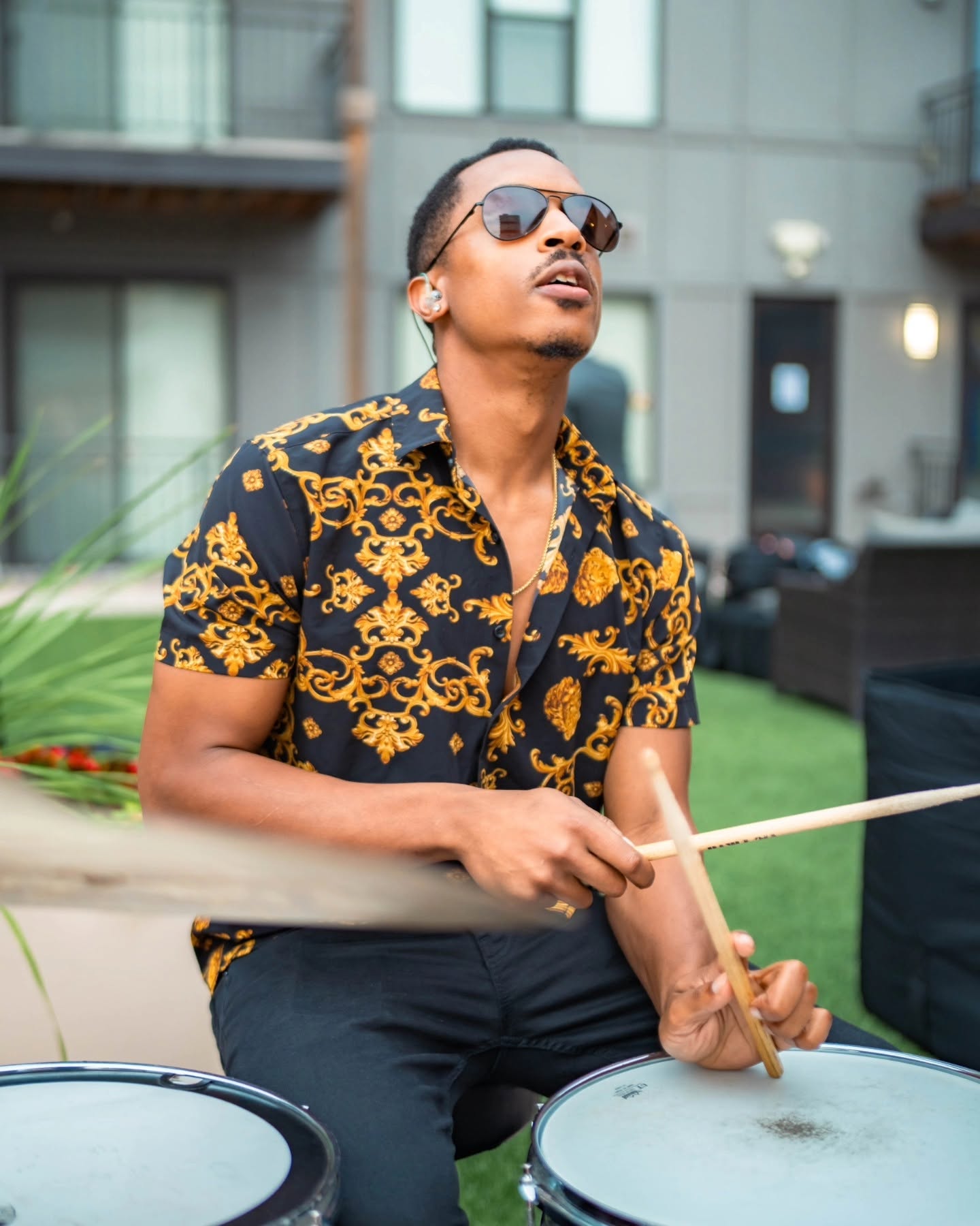 Man playing drums outdoors, wearing sunglasses and a patterned shirt, looking upward with a building in the background.