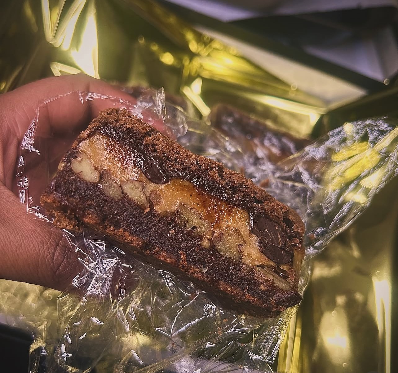 Hand holding a chocolate chip cookie with visible nuts, wrapped in clear plastic, with a shiny gold background.