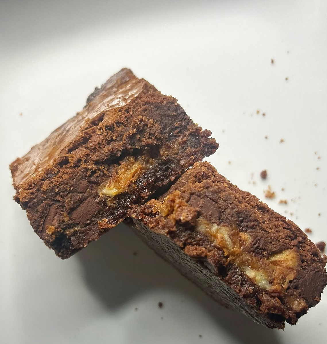 Two chocolate brownies with nuts stacked on a white surface, with crumbs around them.