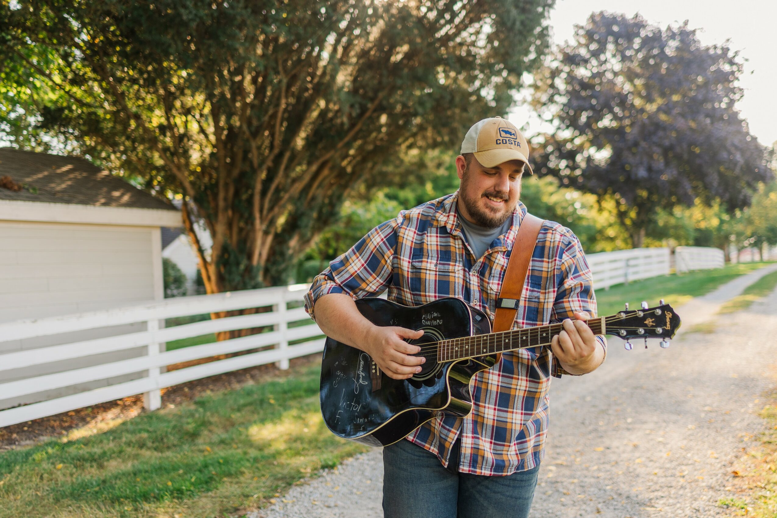 Man playing guitar outdoors near a white fence and trees, wearing a plaid shirt and cap.