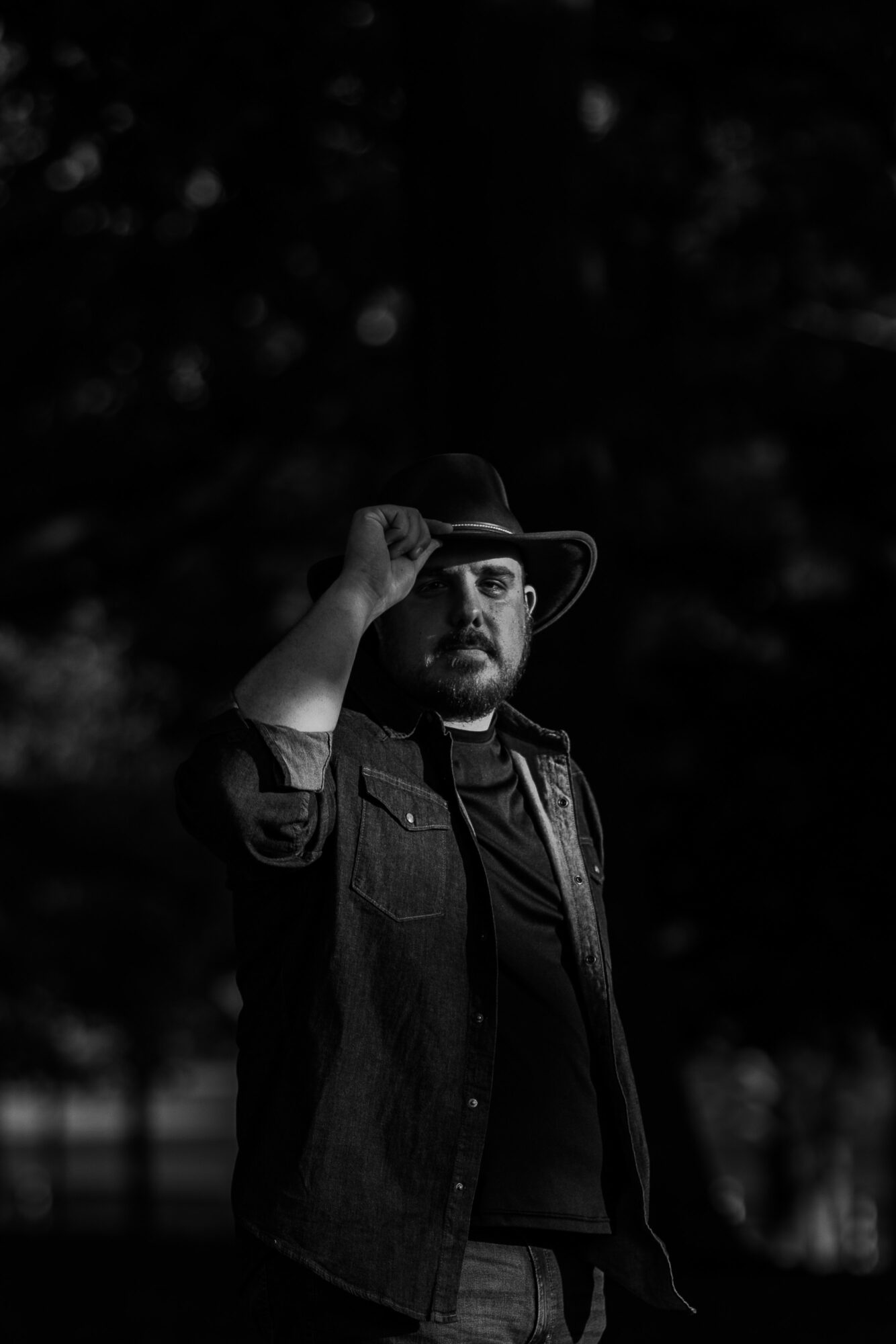 Man wearing a hat and jacket, standing outdoors in a dark, wooded area, looking at the camera.