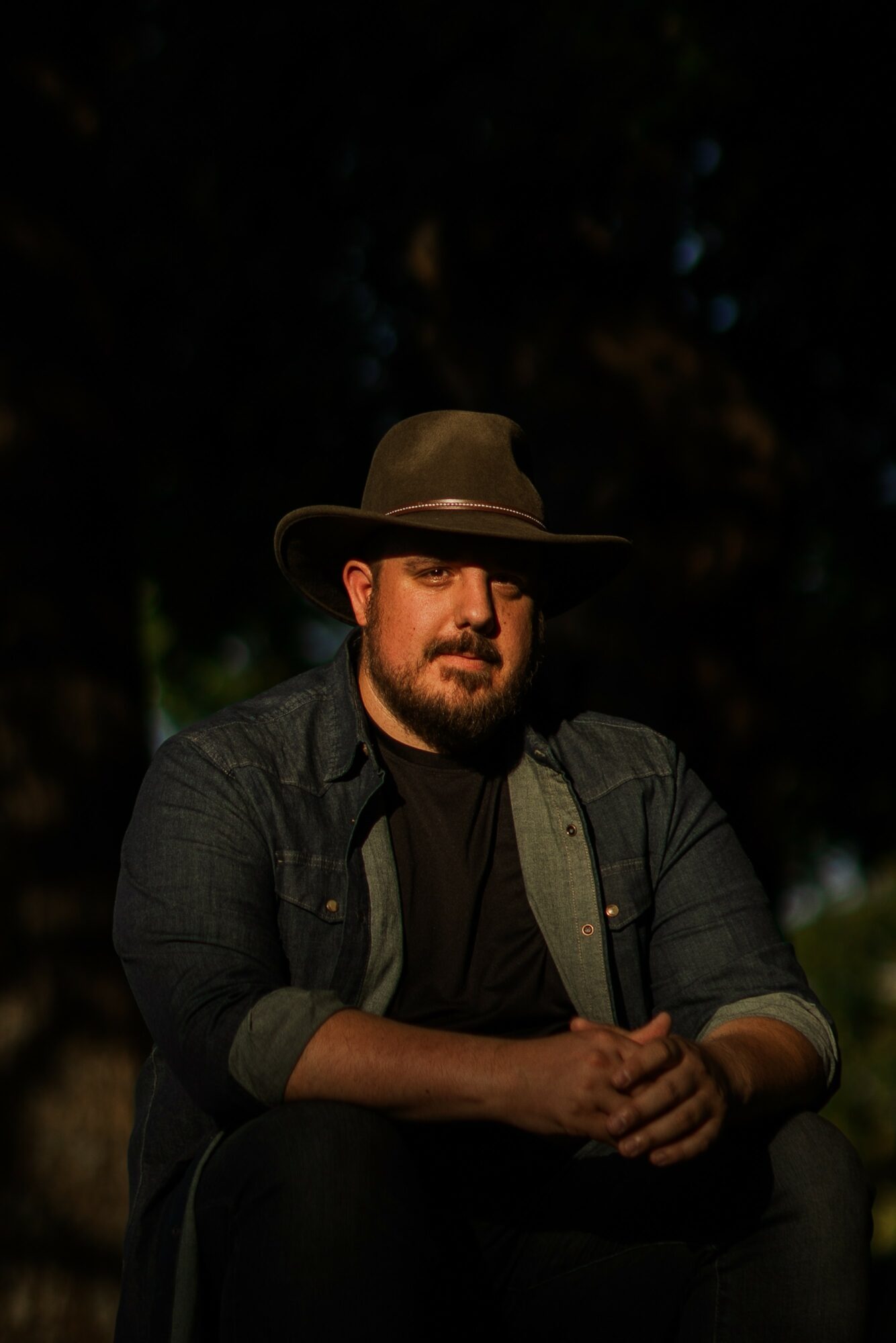 Man with a beard wearing a wide-brimmed hat and dark clothing, sitting outdoors at night.