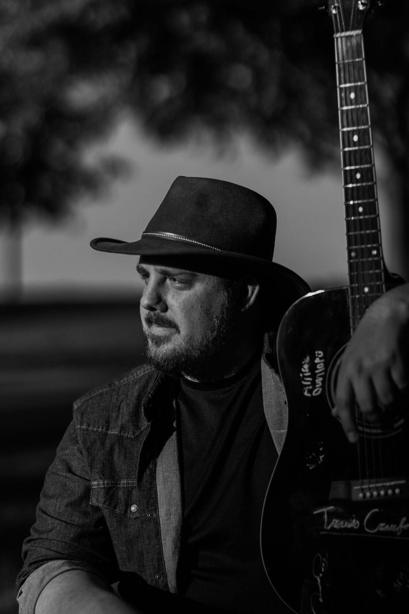 Man wearing a hat and jacket holding an acoustic guitar outdoors, with blurred background.