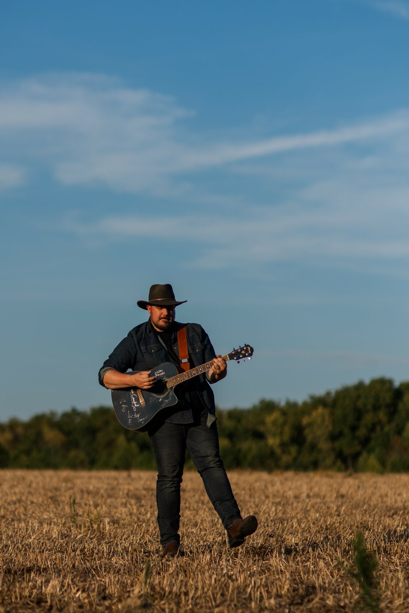 Man wearing a hat playing guitar in an open field under a blue sky with clouds.