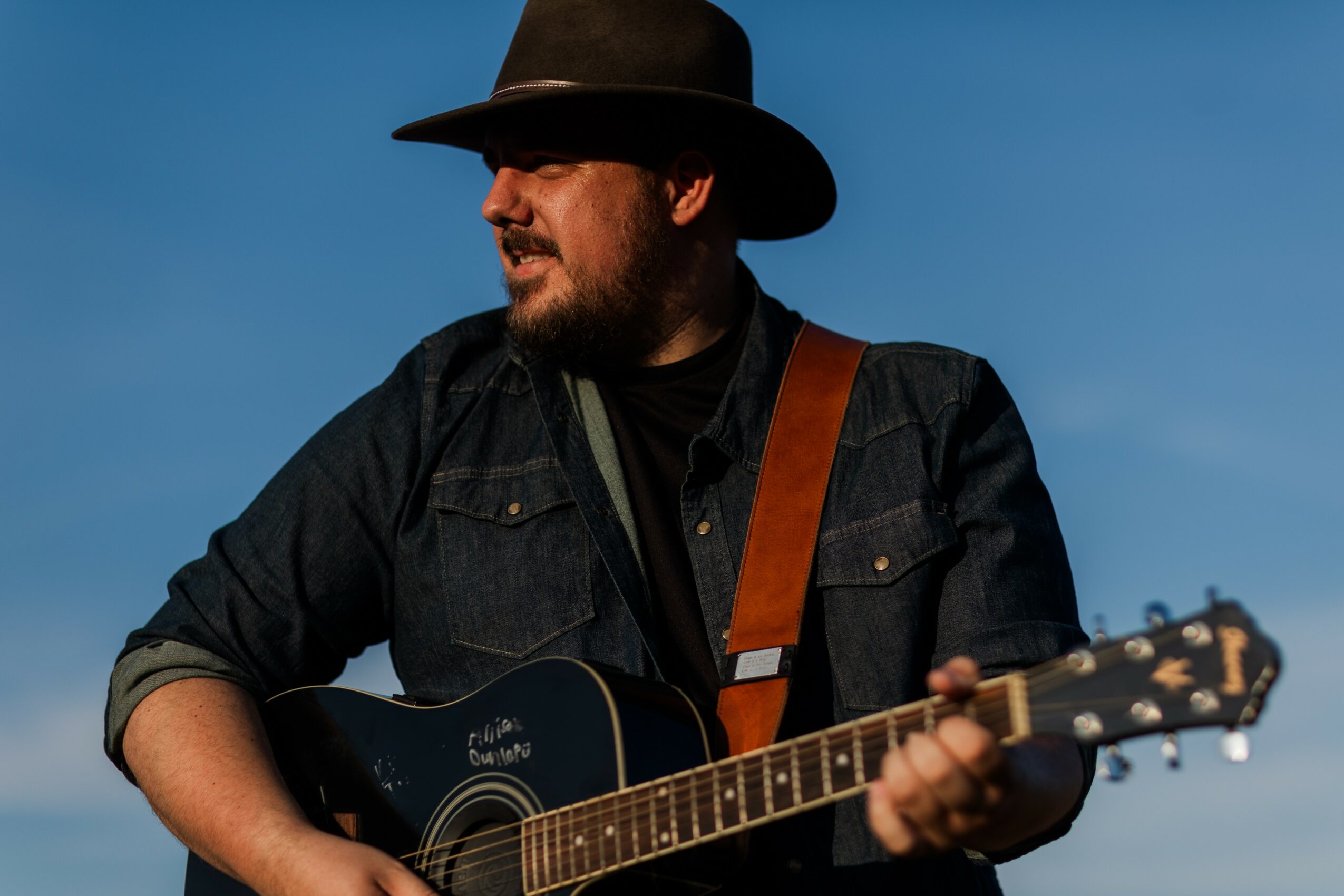 Man wearing a hat playing an acoustic guitar outdoors against a blue sky.