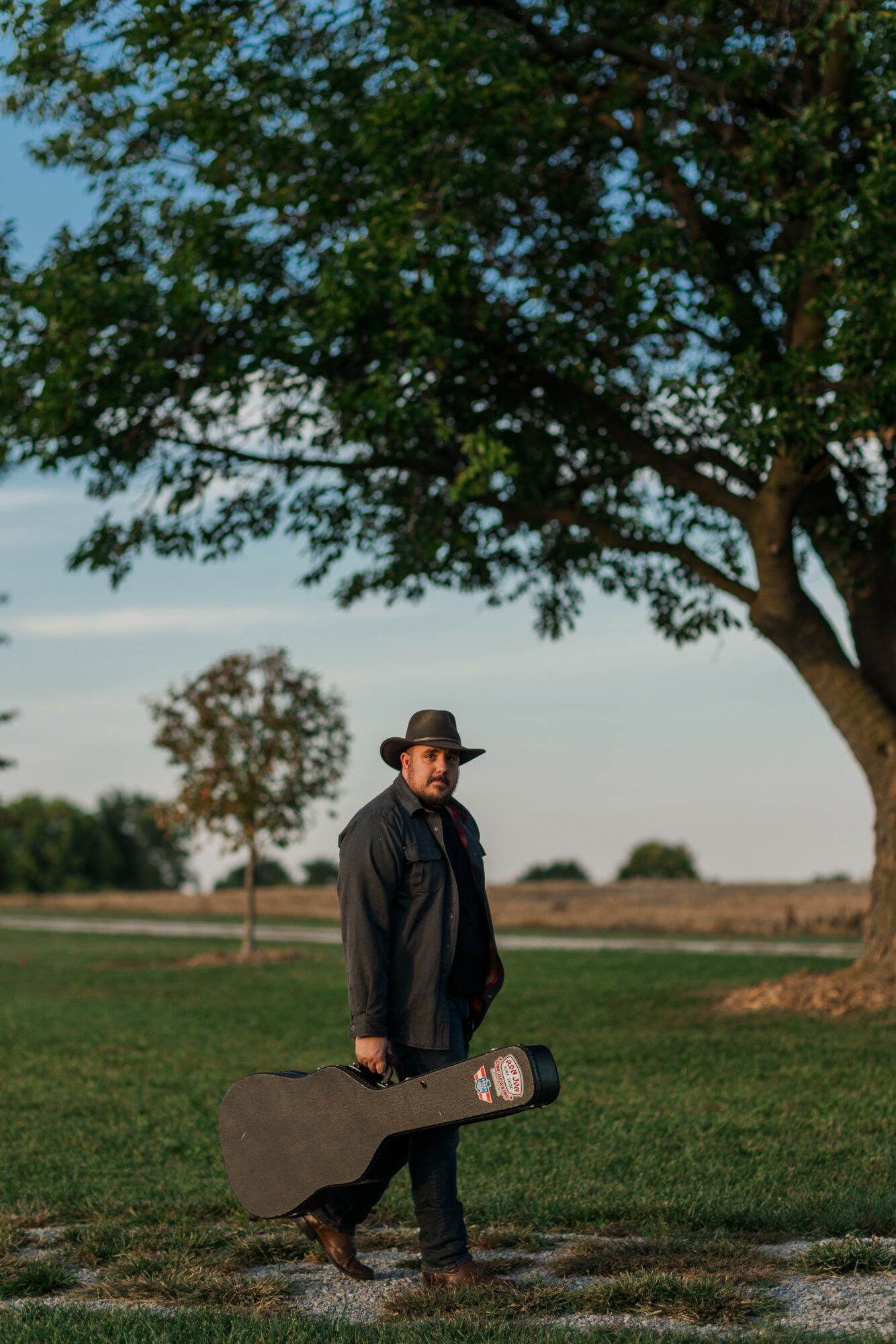 Man standing outdoors holding a guitar case, with trees and open field in background.