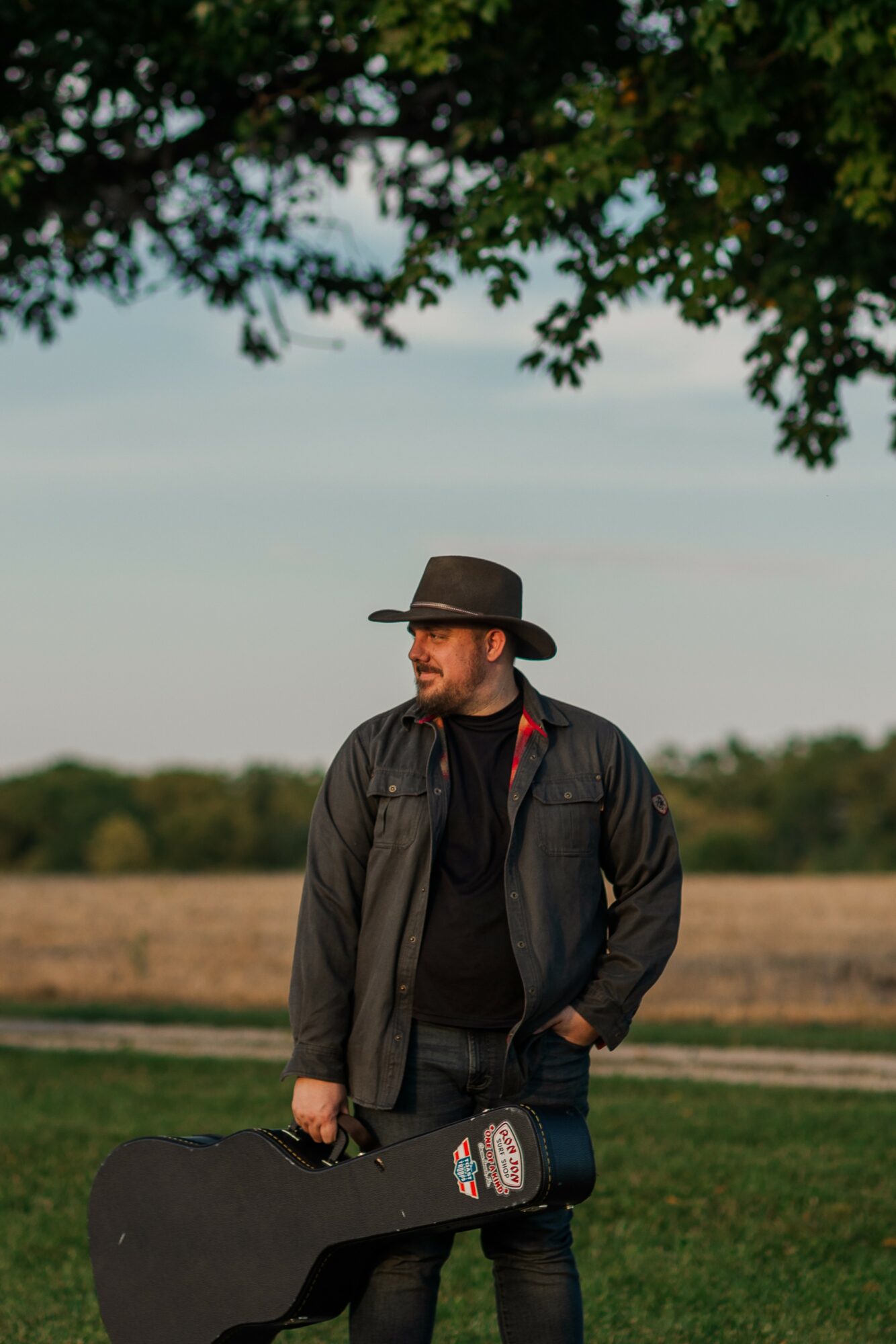 Man wearing a hat and jacket holding a guitar case outdoors under a tree, with fields in the background.