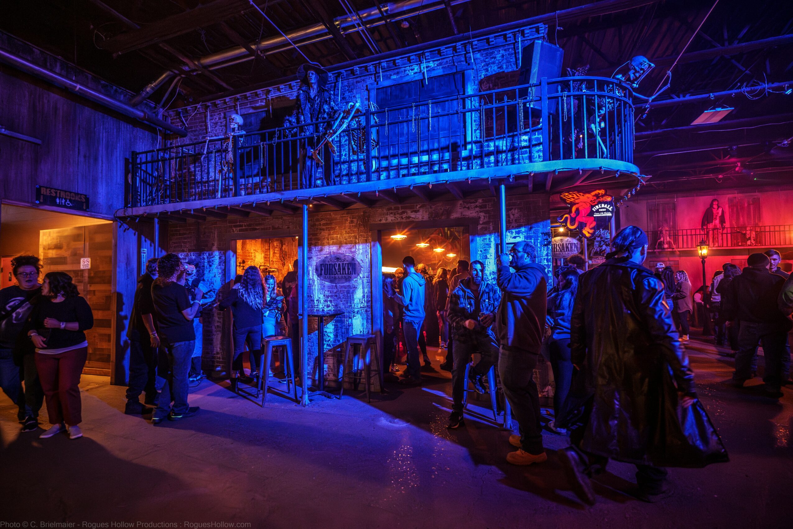 People gather in a dimly lit space with blue and red lighting, featuring a large metal structure above and a crowd below.