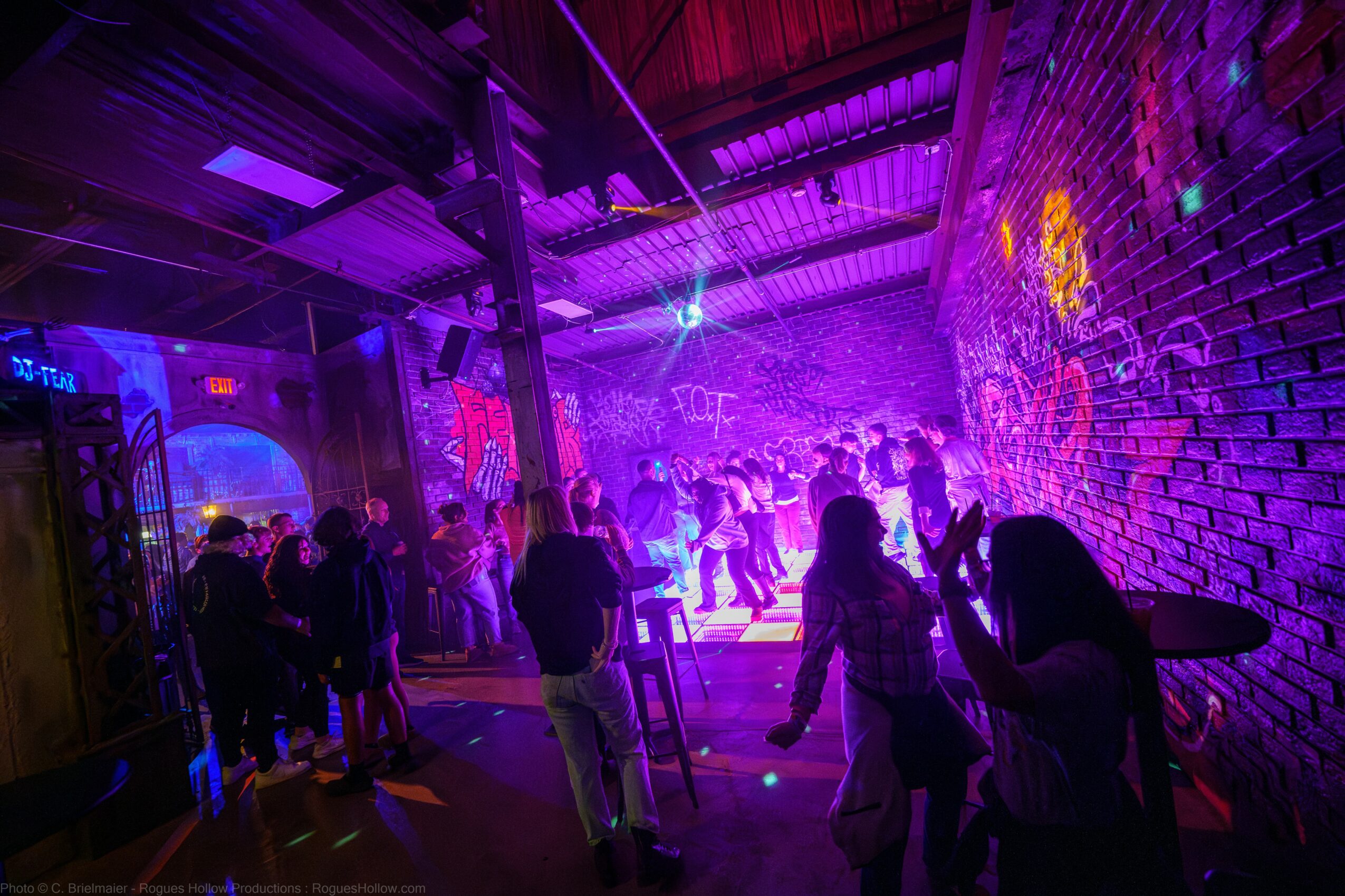 People dancing and socializing in a colorful, neon-lit indoor space with graffiti-covered brick walls.