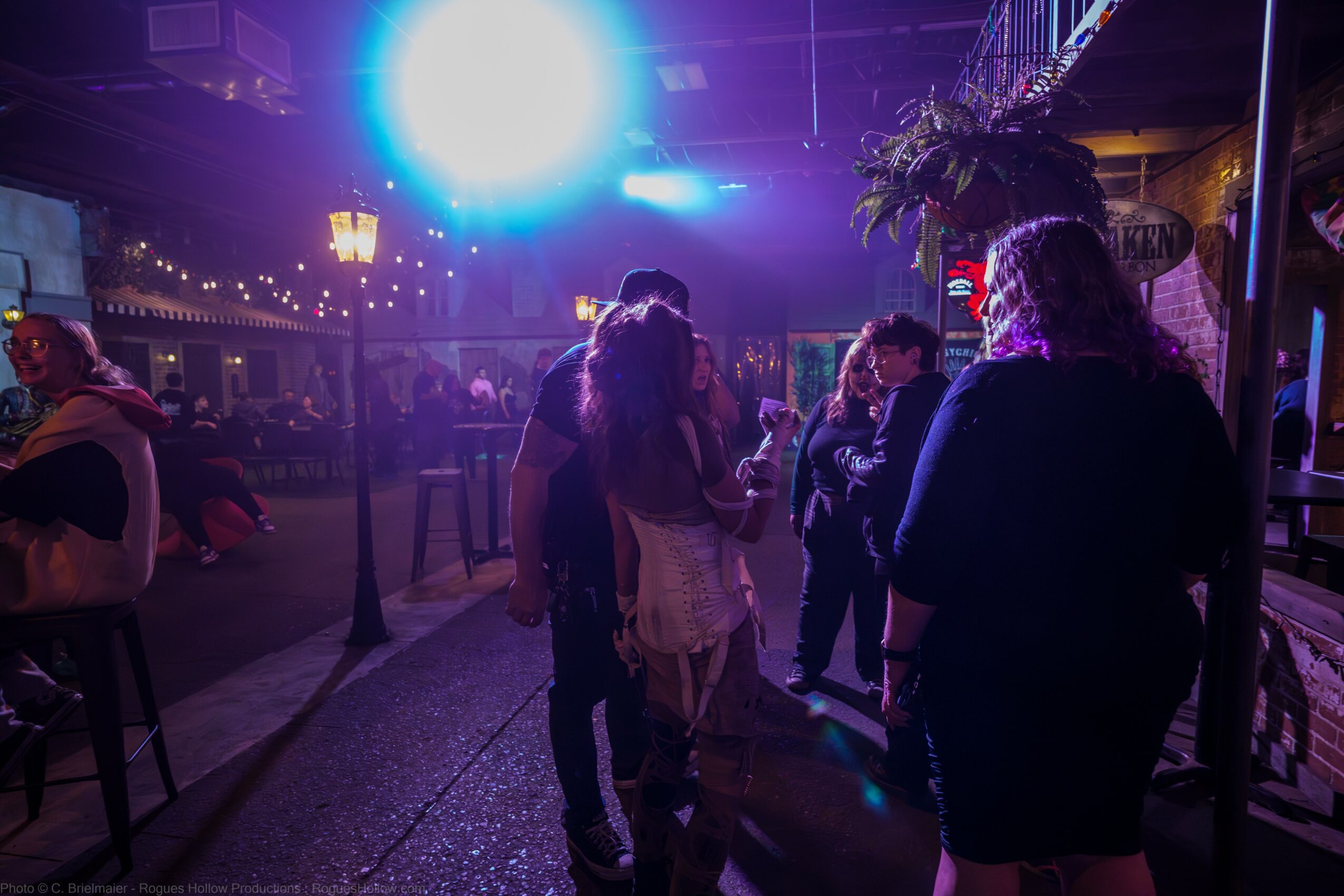 People dancing and socializing in a dimly lit venue with colorful lights and a large bright light in the background.