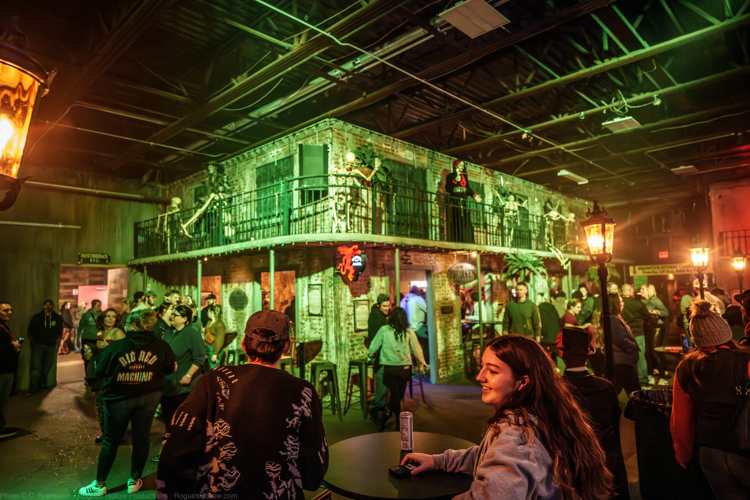 People gather inside a dimly lit venue with green lighting, a two-story structure, and festive decorations, including wreaths.