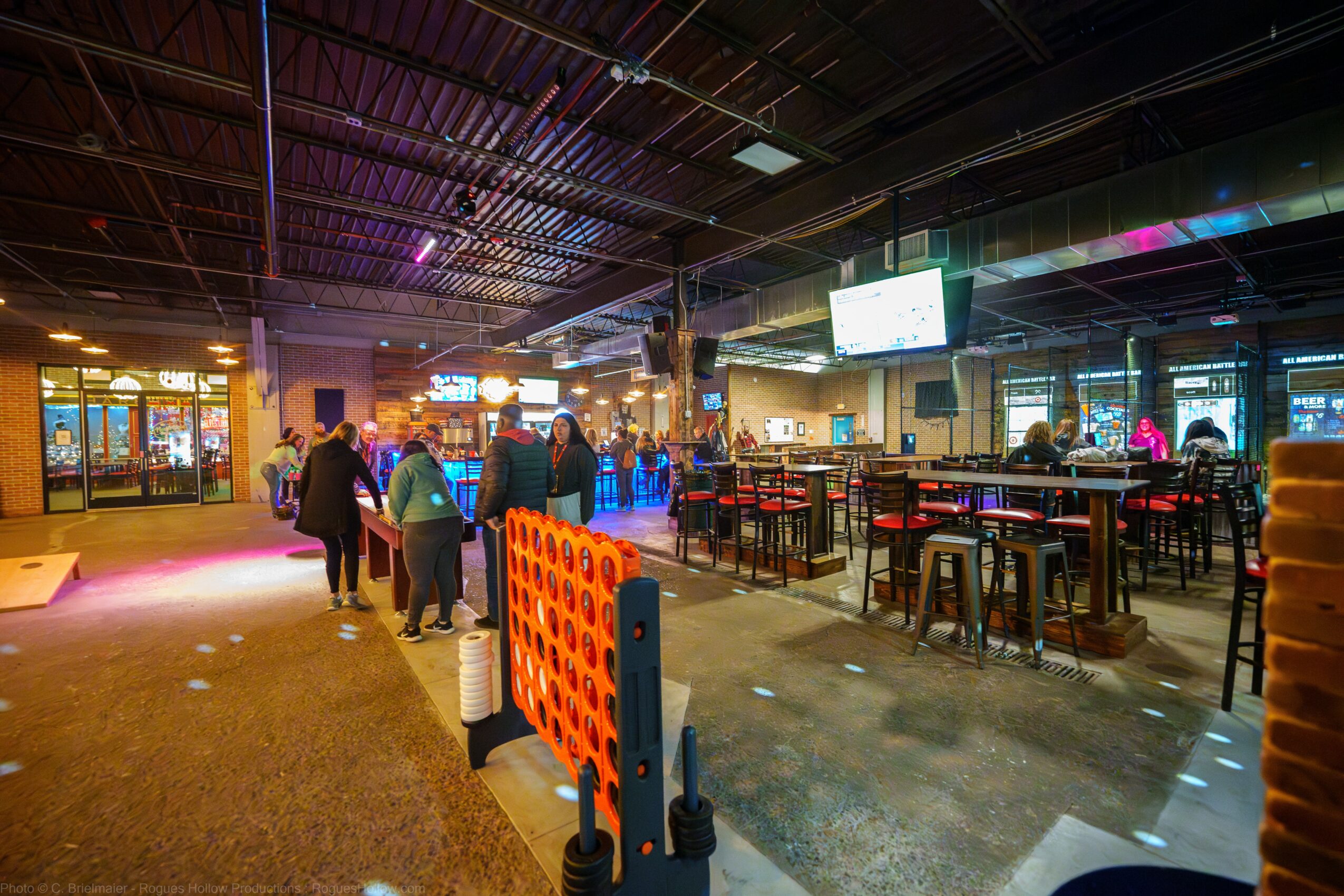 Indoor entertainment venue with seating, tables, and a bar area, illuminated by colorful lighting, with people socializing.