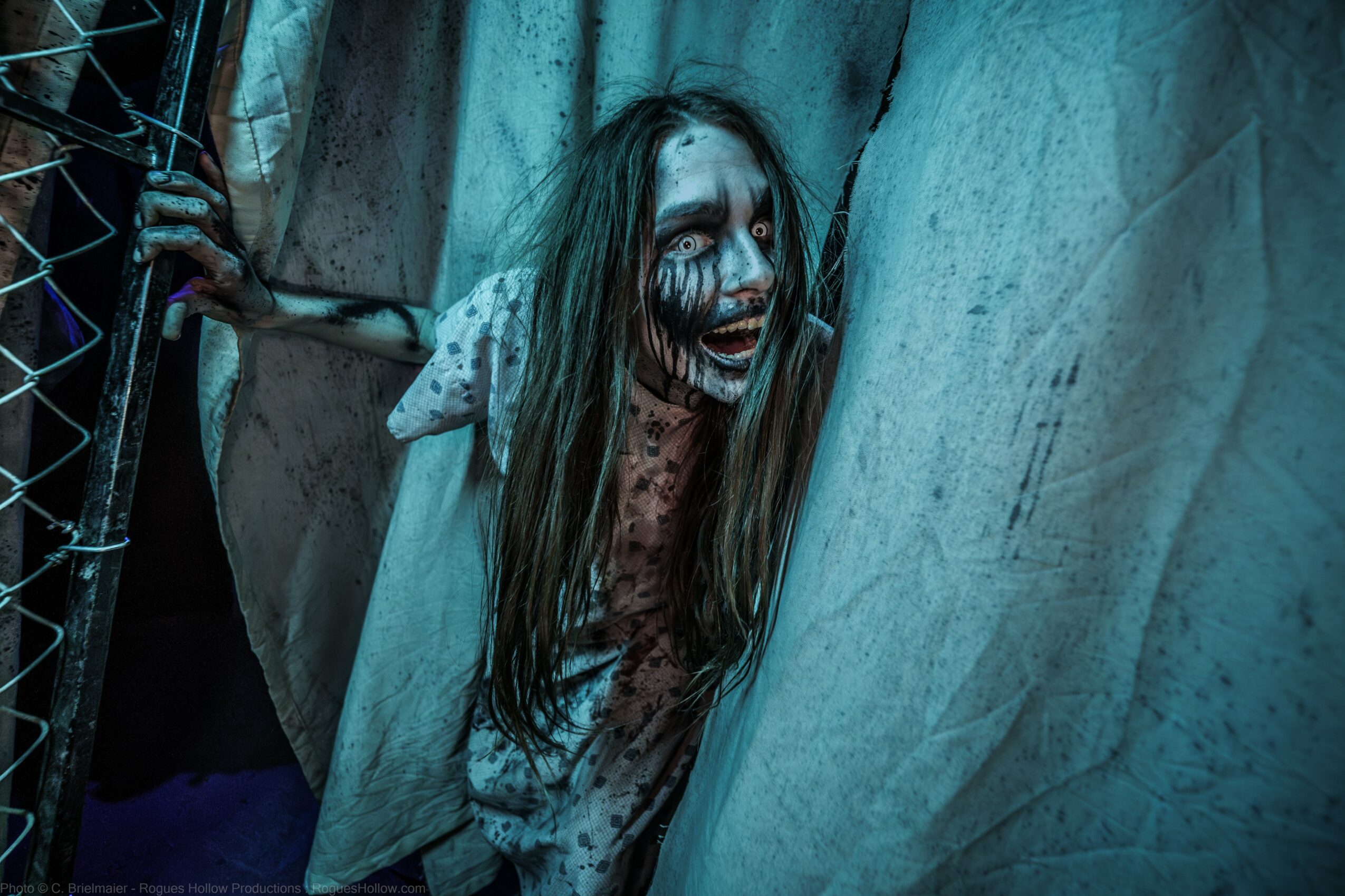 Person with face painted as a creepy clown, peeking from behind a curtain, with a chain-link fence nearby.