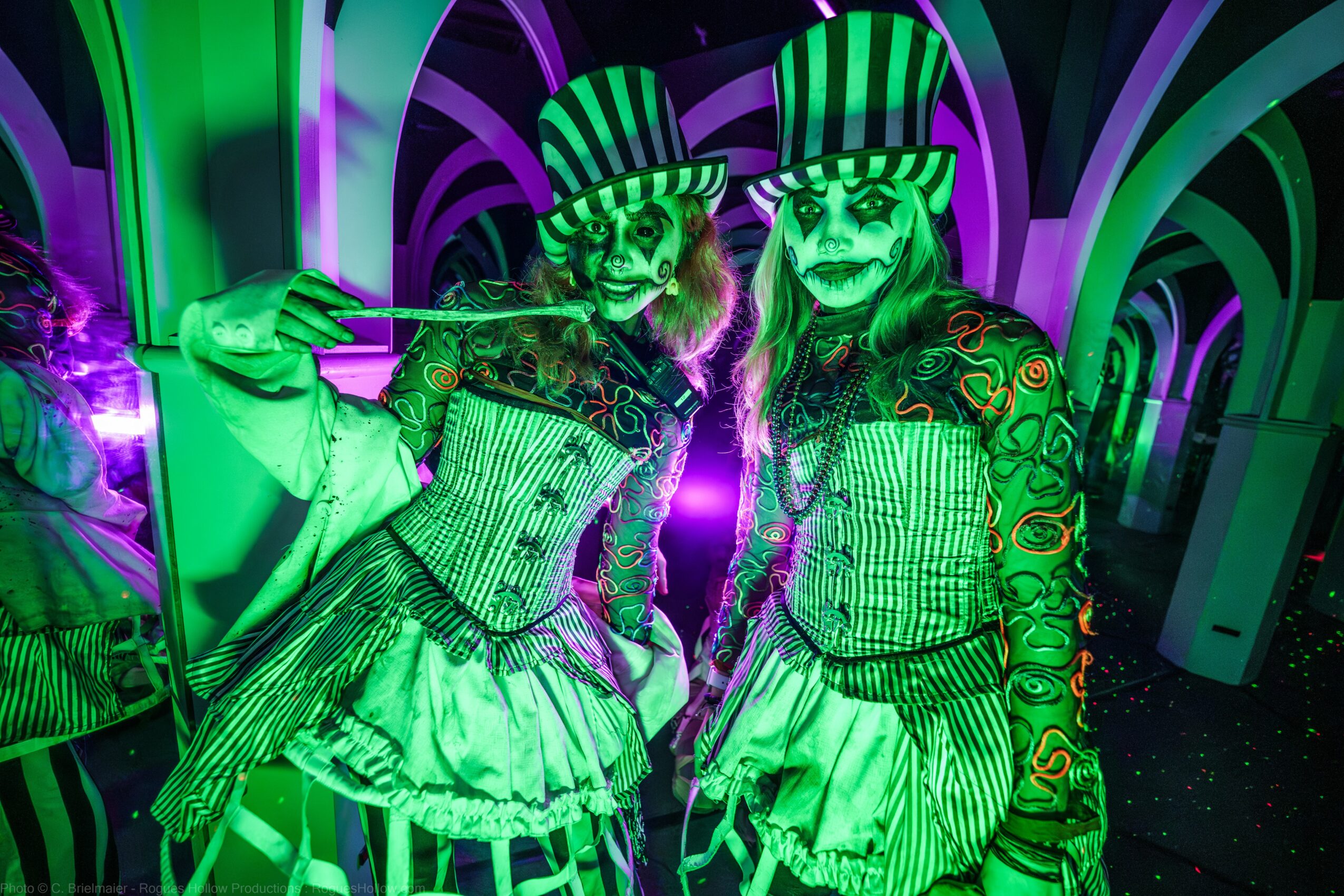 Two performers in green costumes and striped hats with painted faces in a colorful, neon-lit setting.