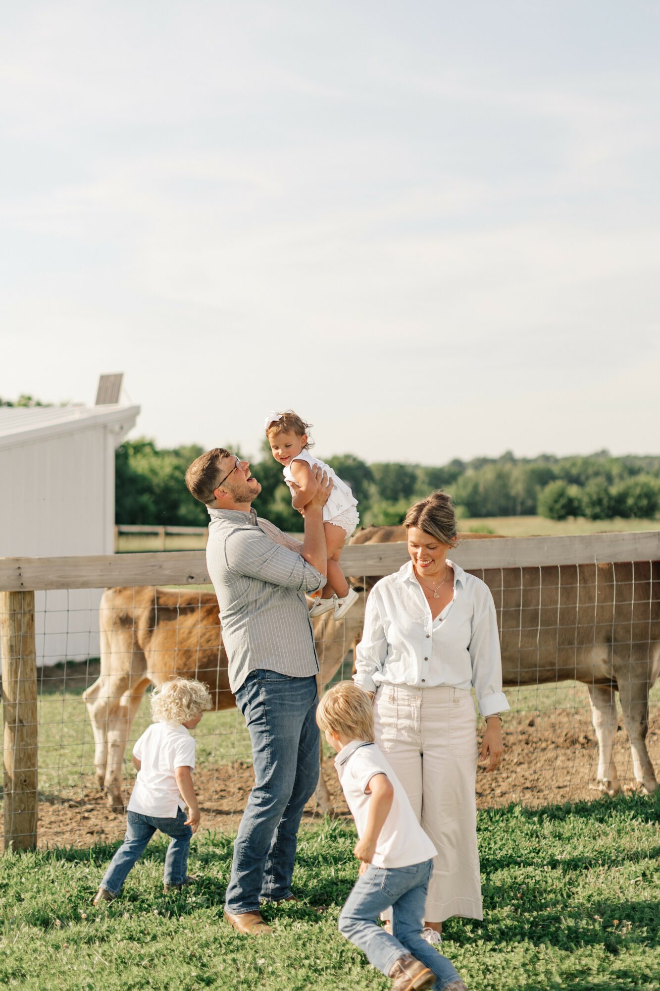 Family outdoors with two children, a man lifting a child, woman standing nearby, cows in background, sunny day.