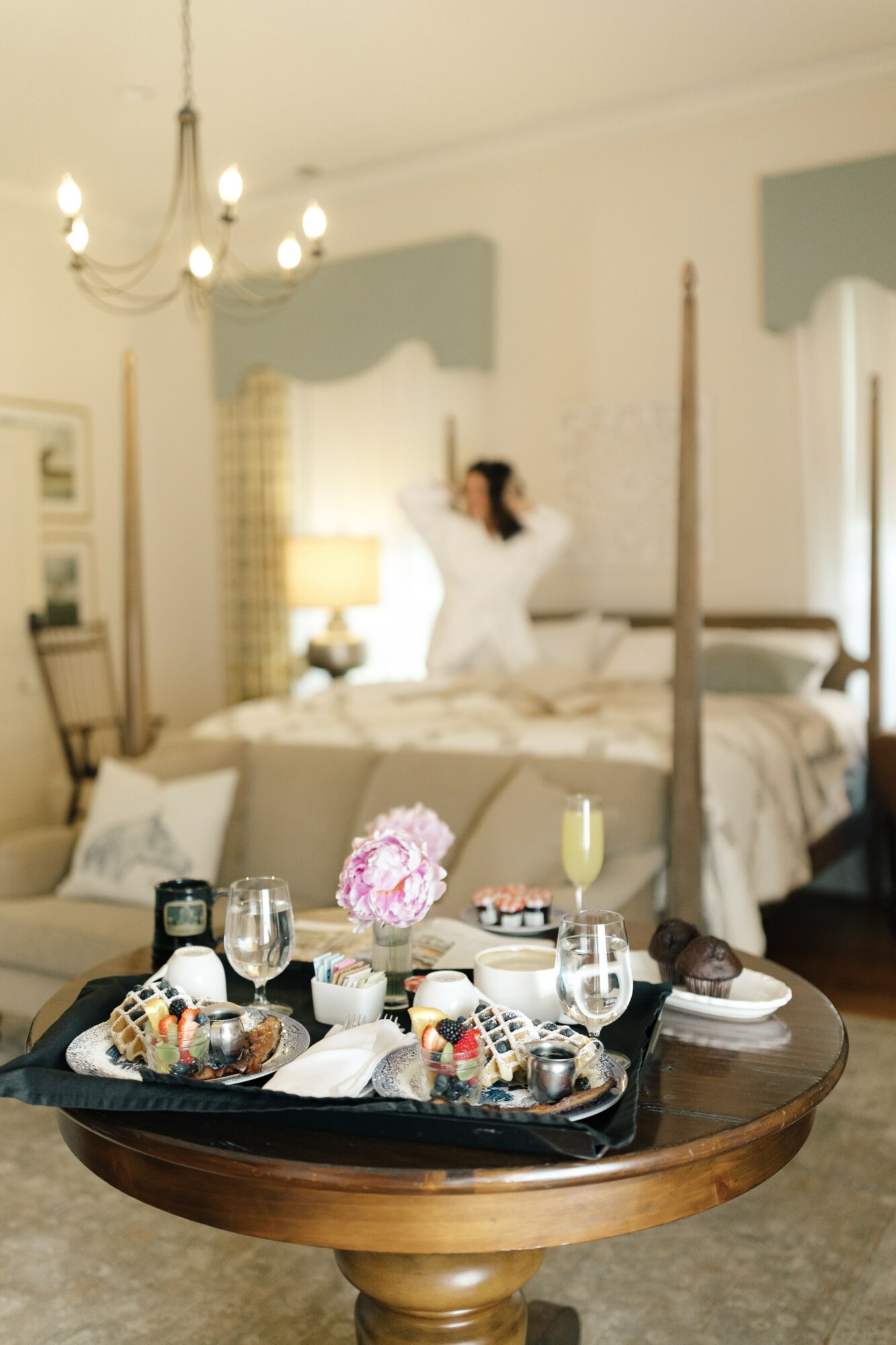 A bedroom with a woman in white dress standing near the bed, a tray with food and drinks on a round table in foreground.