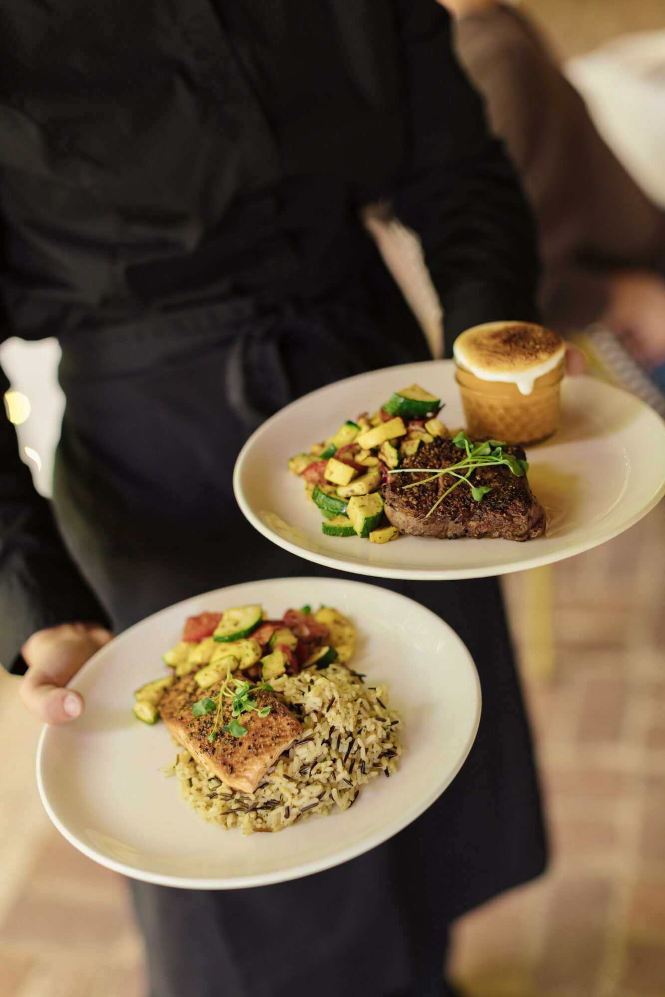 Person holding two plates with food, one with a burger and salad, the other with rice and a piece of meat, in a restaurant setting.
