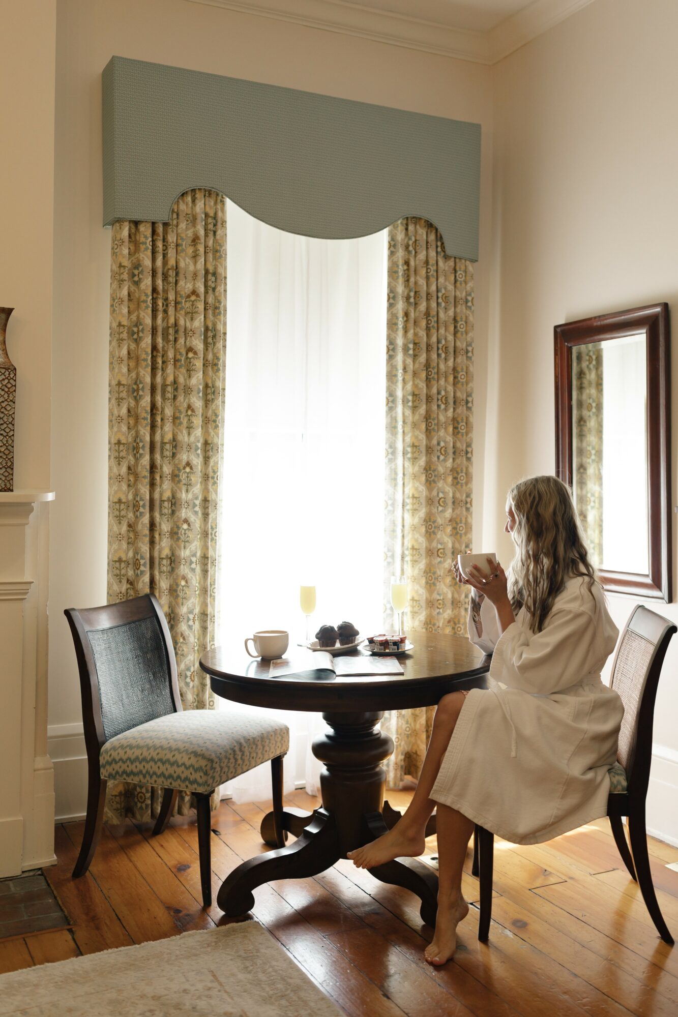 Woman in a white robe sitting at a round table with a cup, in a room with large window, curtains, and mirror.
