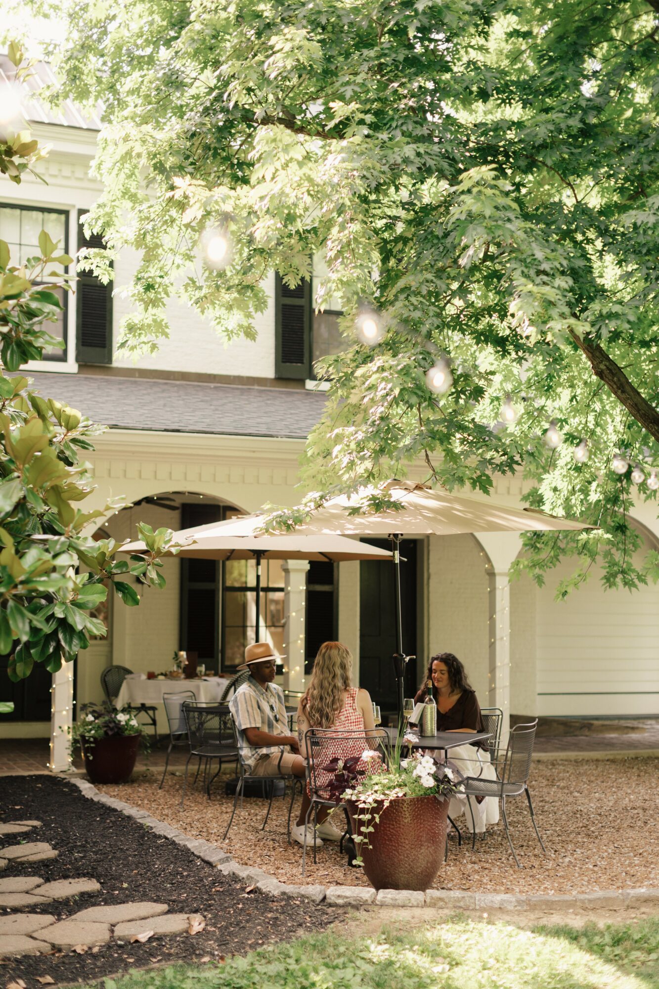 Three people sit at a table outside a house with a porch, surrounded by trees and potted plants, on a sunny day.