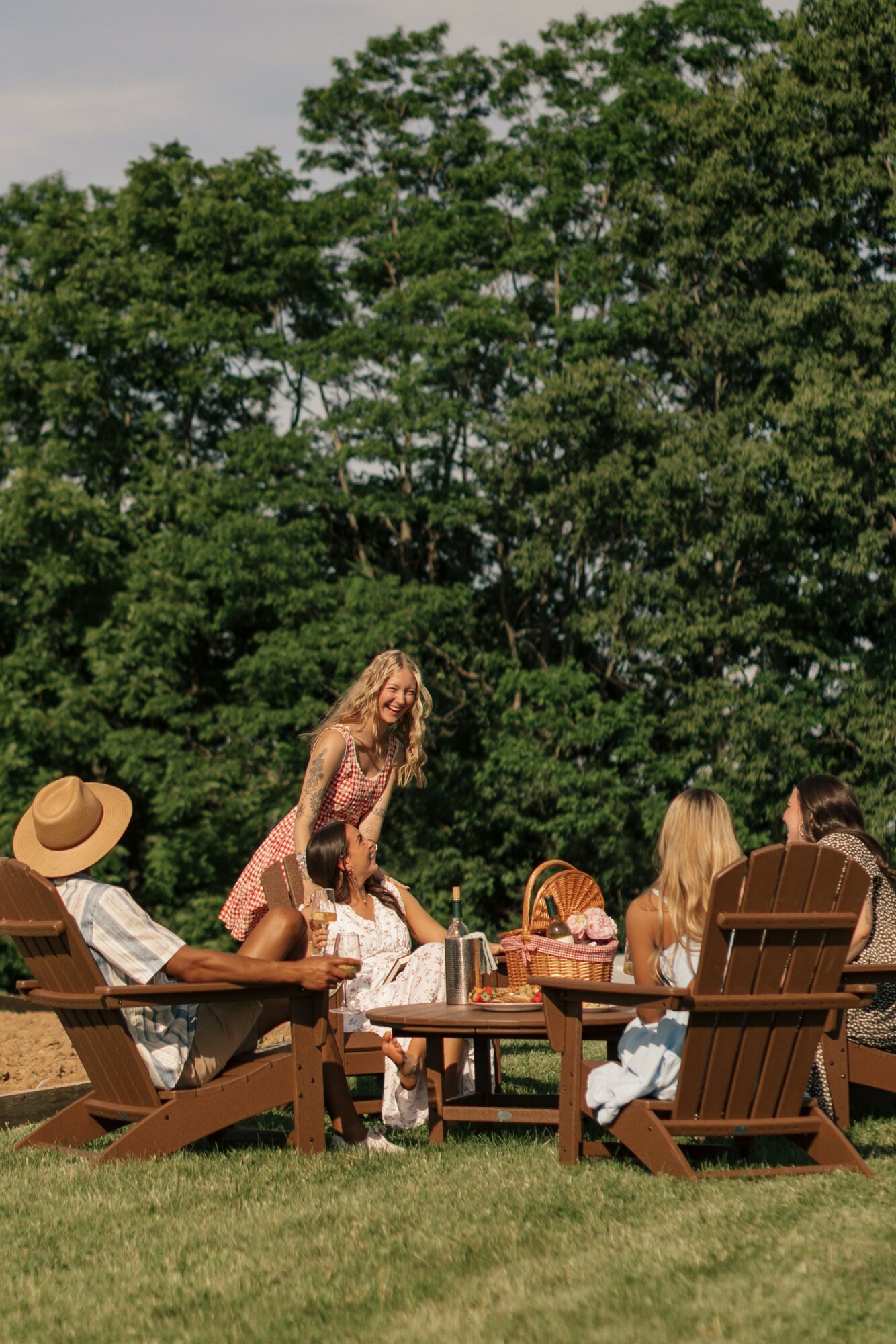 Group of people outdoors with trees in background, sitting and standing around a table, enjoying a sunny day.