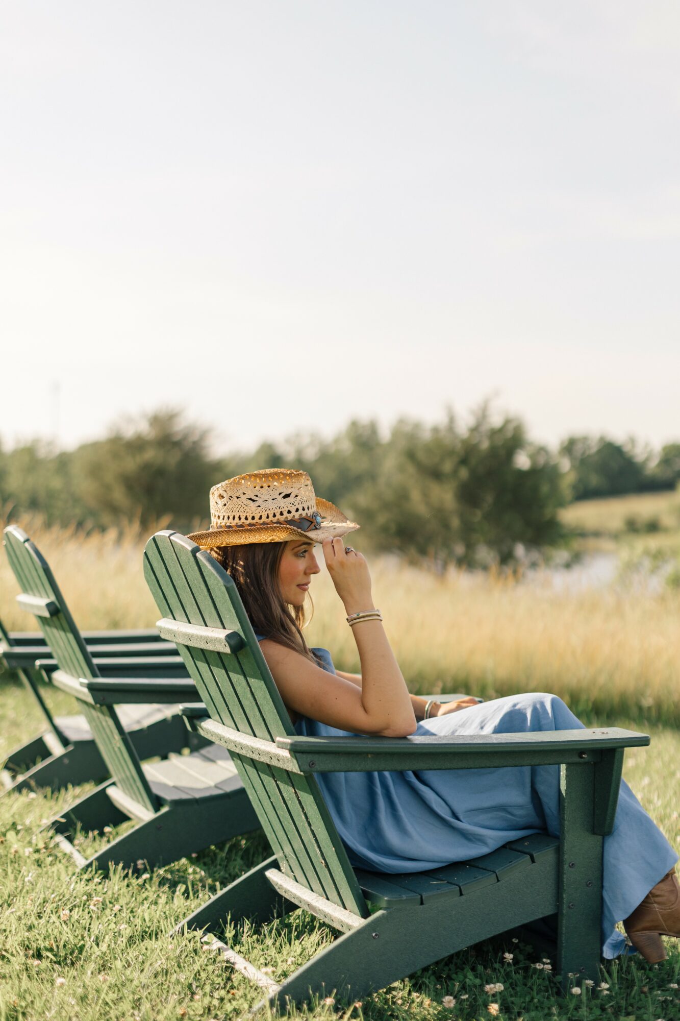 Woman sitting on a green chair outdoors, wearing a straw hat and blue dress, with a field and trees in background.