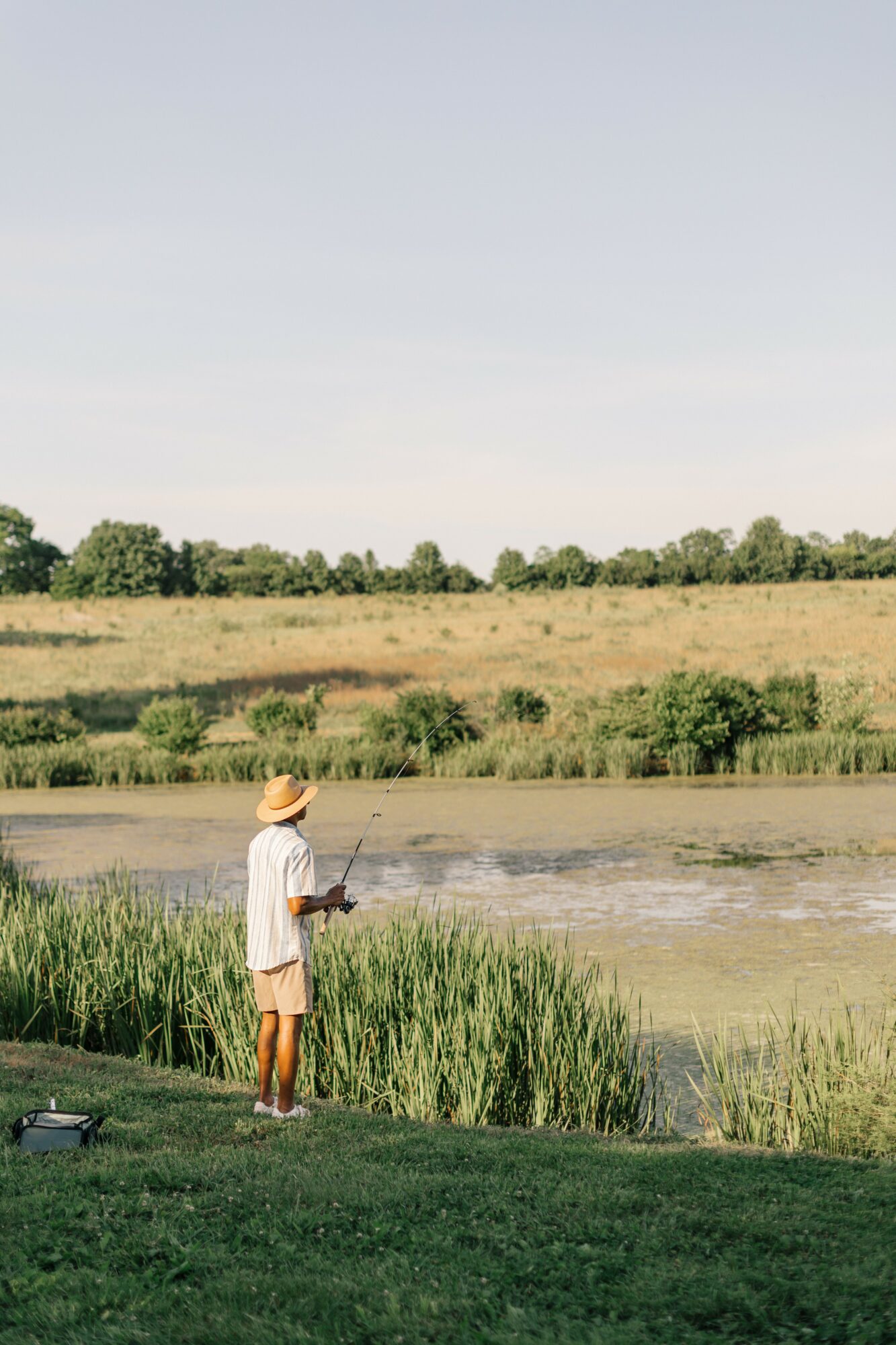 Child fishing by a pond with grassy banks and a field in the background under a clear sky.