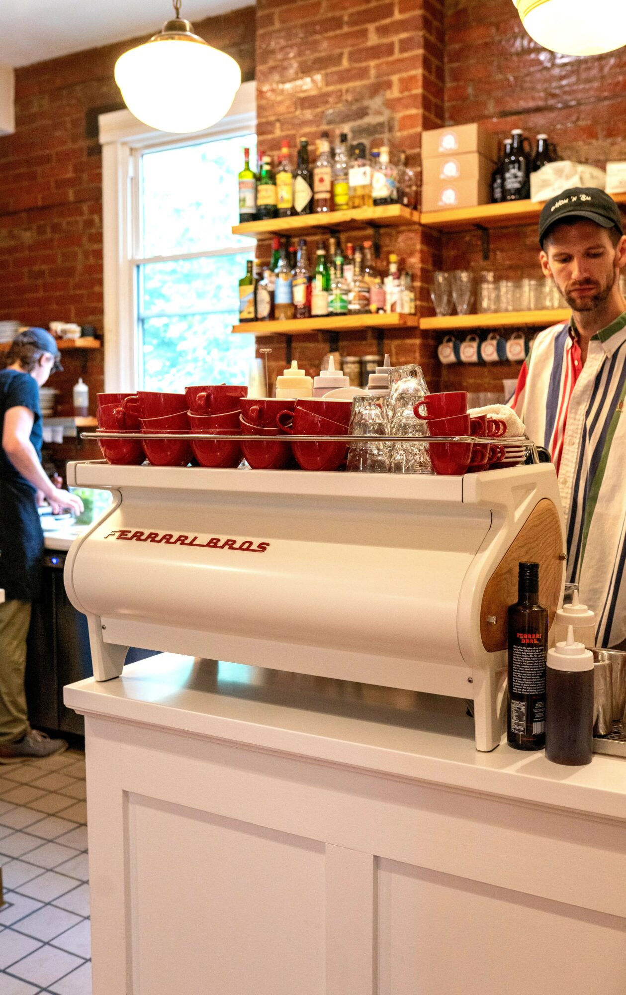Barista behind counter with red cups and coffee machine, shelves with bottles and jars, brick wall background, hanging lights.