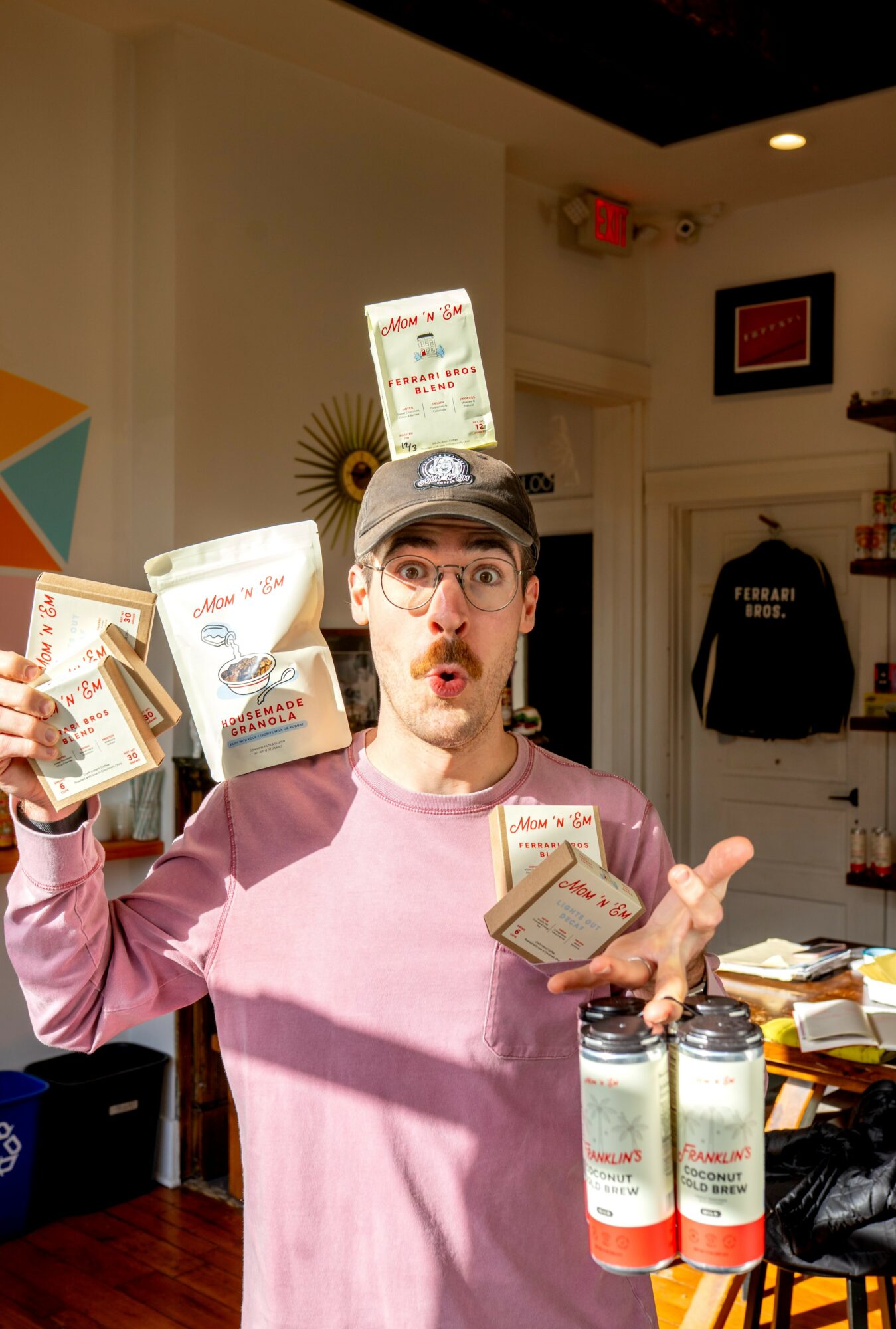 Man with glasses and a cap holding snack packages, pointing with one hand, in a room with shelves and wall decorations.