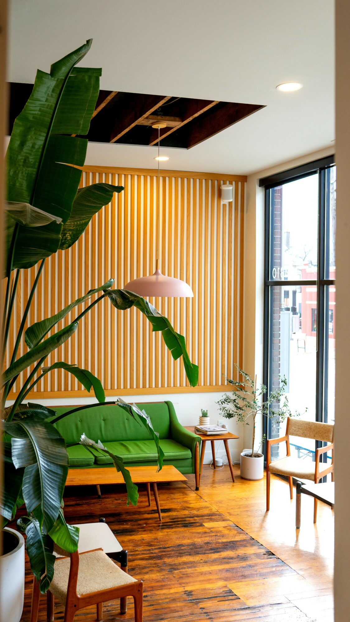 Interior living space with green sofa, wooden floor, large window, and yellow vertical blinds, decorated with plants and furniture.