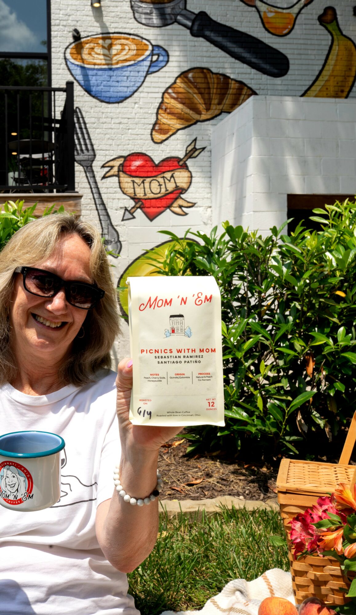 Woman holding a takeout bag outdoors, smiling, with plants and a mural in the background.
