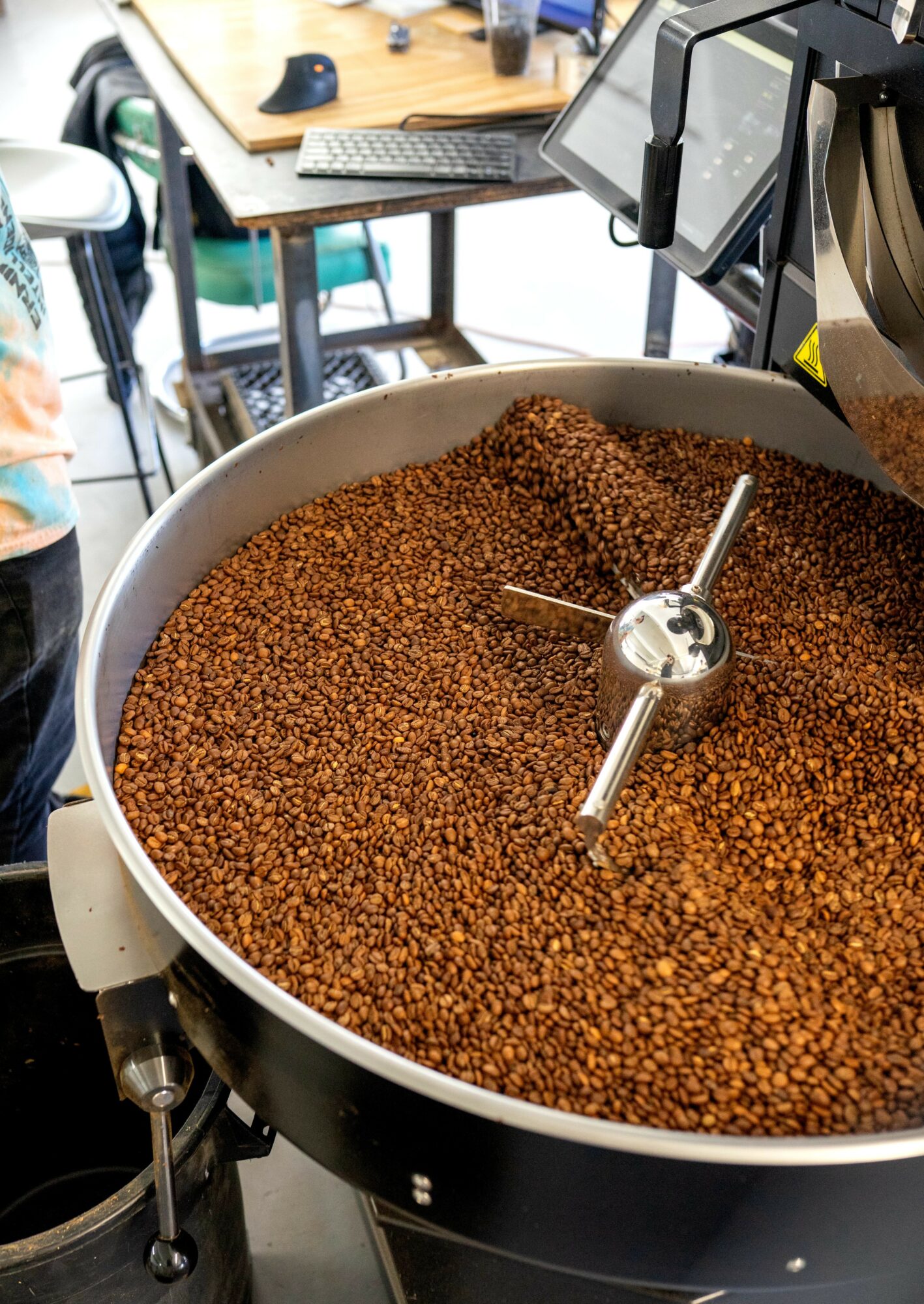 Large container filled with roasted coffee beans, with a metal paddle inside, in a workspace setting.