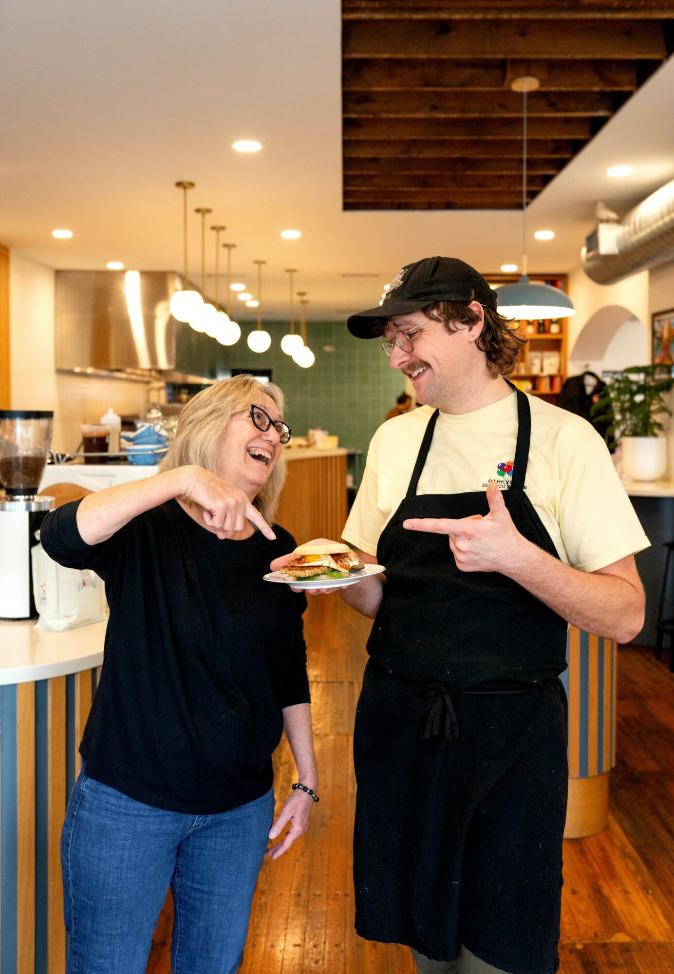 Two people, a woman and a man, smiling and talking in a restaurant kitchen, woman holding a plate with food.
