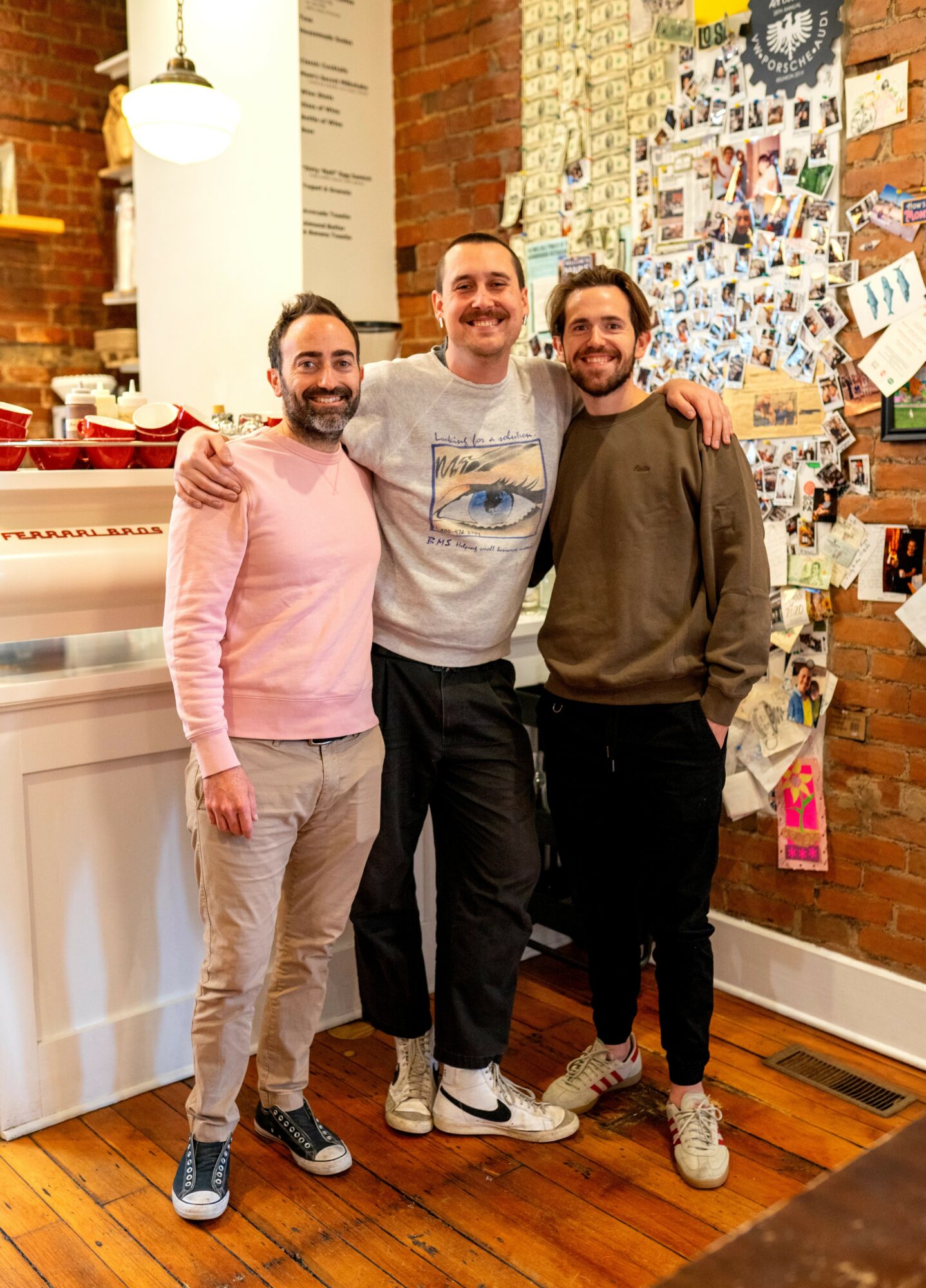 Three men stand together indoors, smiling, with arms around each other, in front of a brick wall decorated with photos and notes.