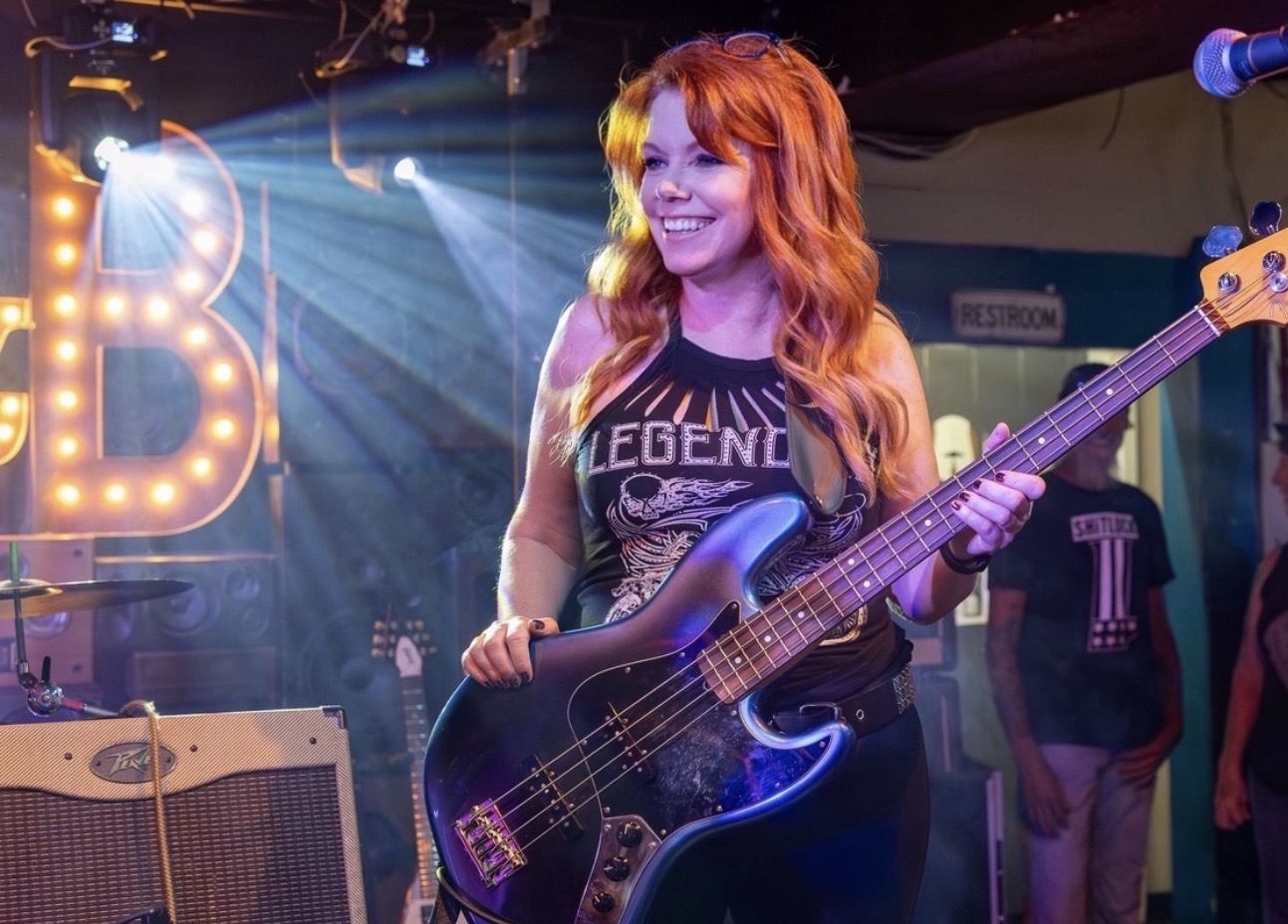 Red-haired woman playing a bass guitar on stage, smiling, with stage lights and equipment in background.