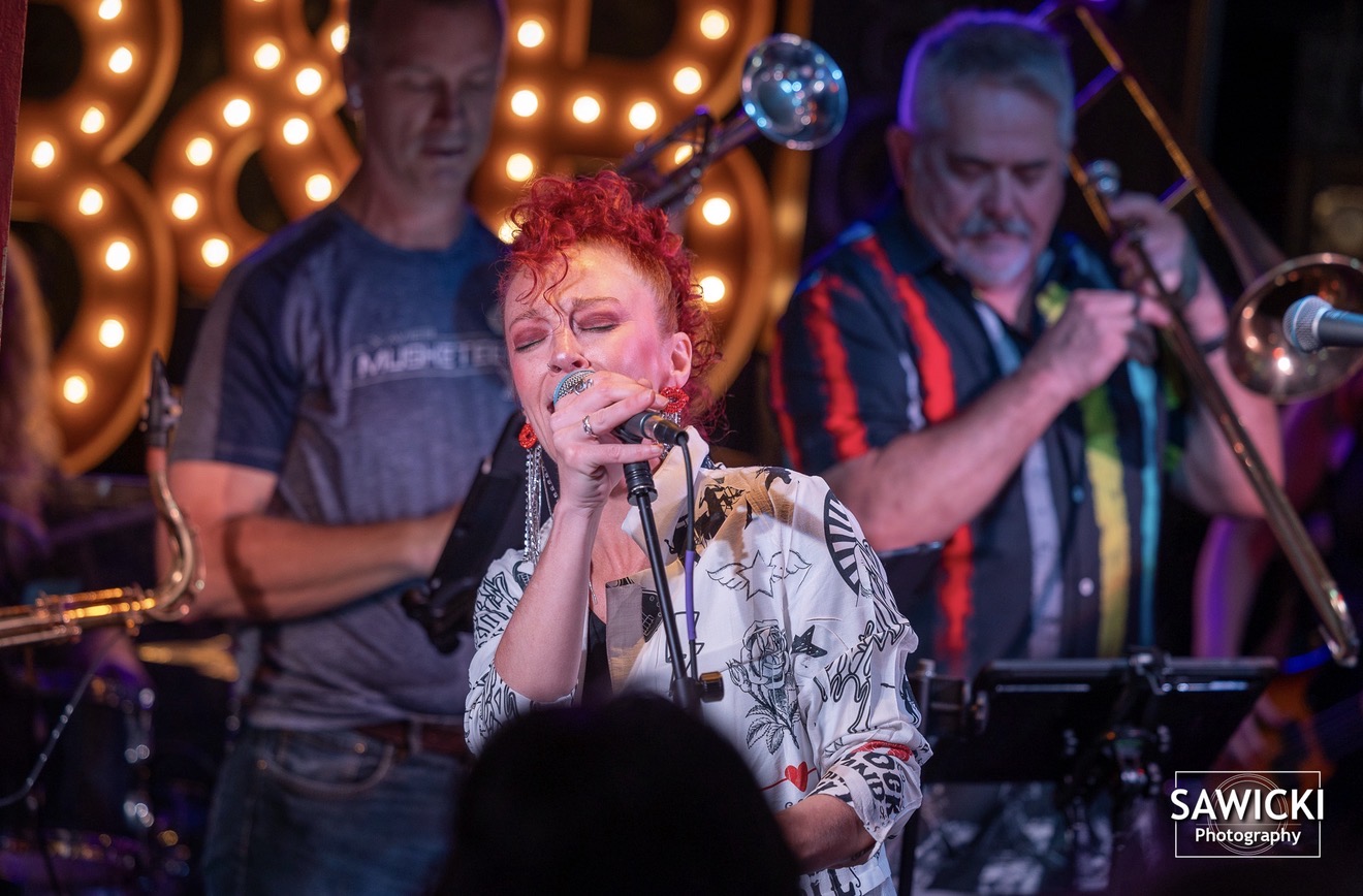 A woman singing into a microphone with two men playing instruments behind her, stage lights in background.
