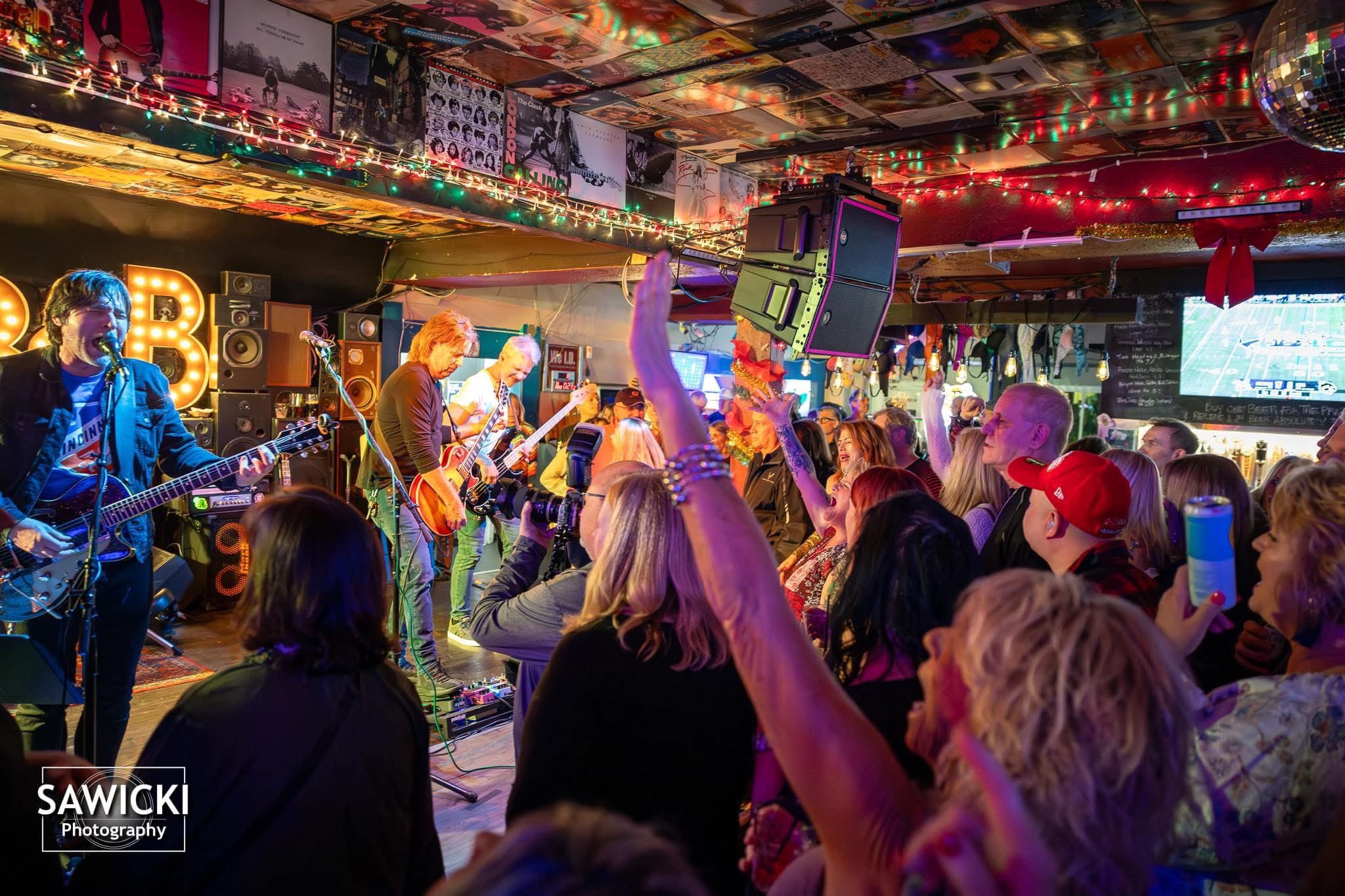 Band performs on stage with audience raising hands and taking photos in a lively indoor venue.