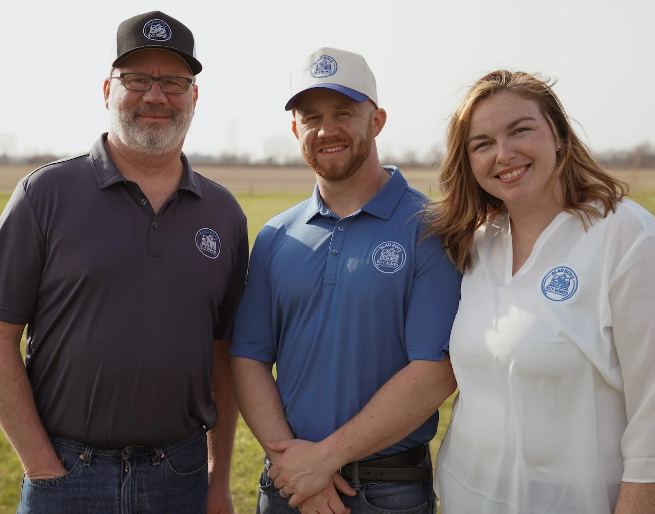 Three people outdoors, smiling, wearing polo shirts and caps with logos, standing on grass field.