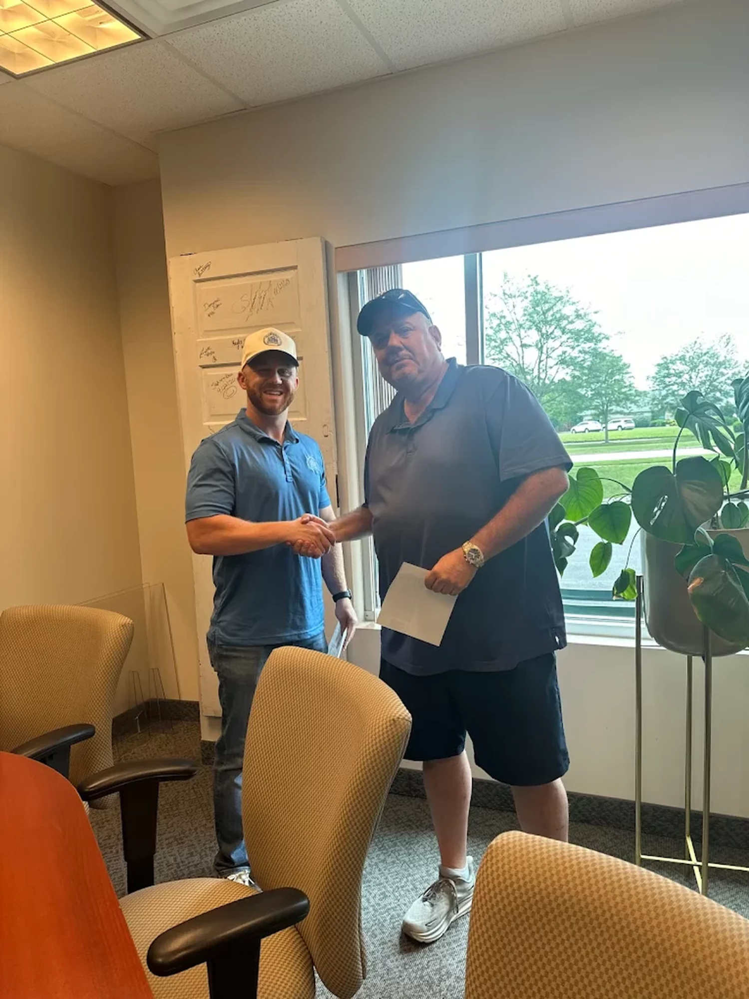 Two men shaking hands inside a room near a window with trees outside, one holding a paper, both smiling.